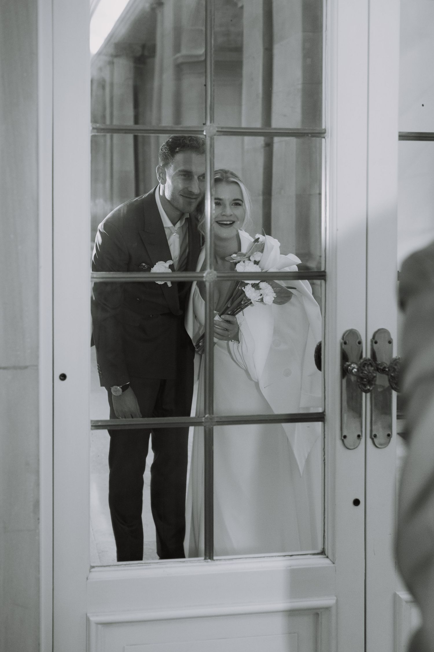 A couple peers through a glass door window on their wedding day, creating a romantic framed portrait.