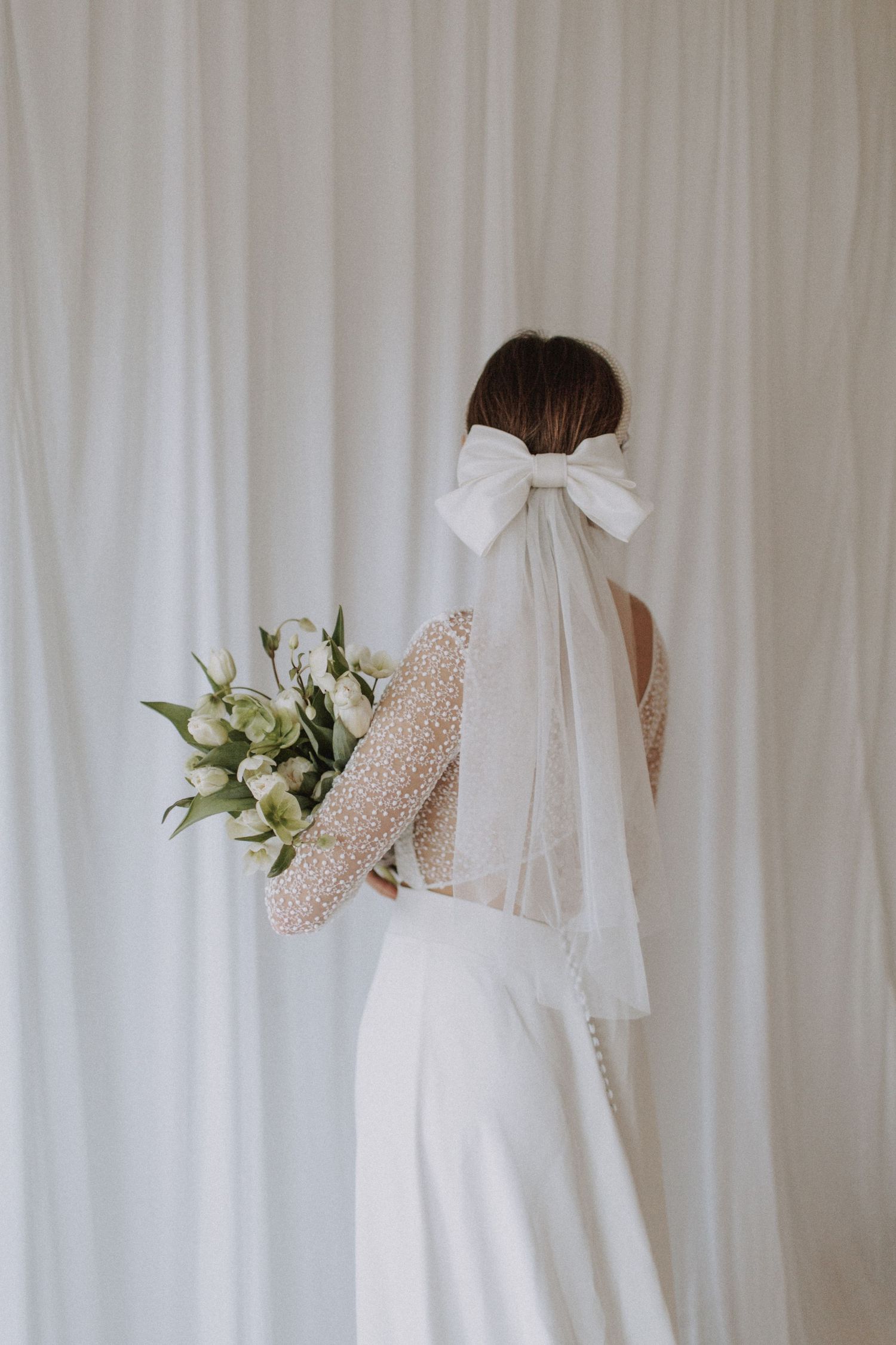 Elegant white wedding dress with bow detail and floral accents against white curtain.