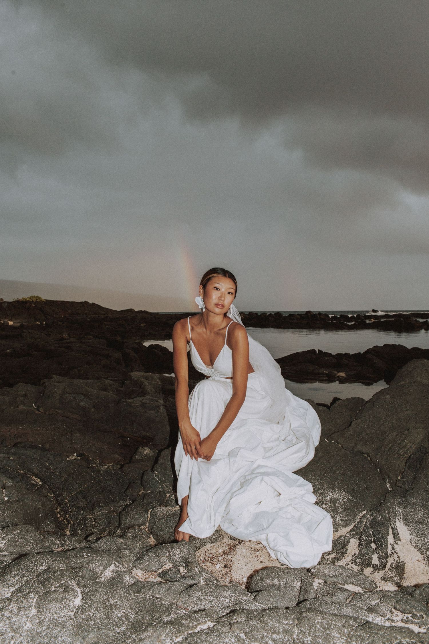 Dramatic bridal portrait on rocky coastline with stormy sky and rainbow.