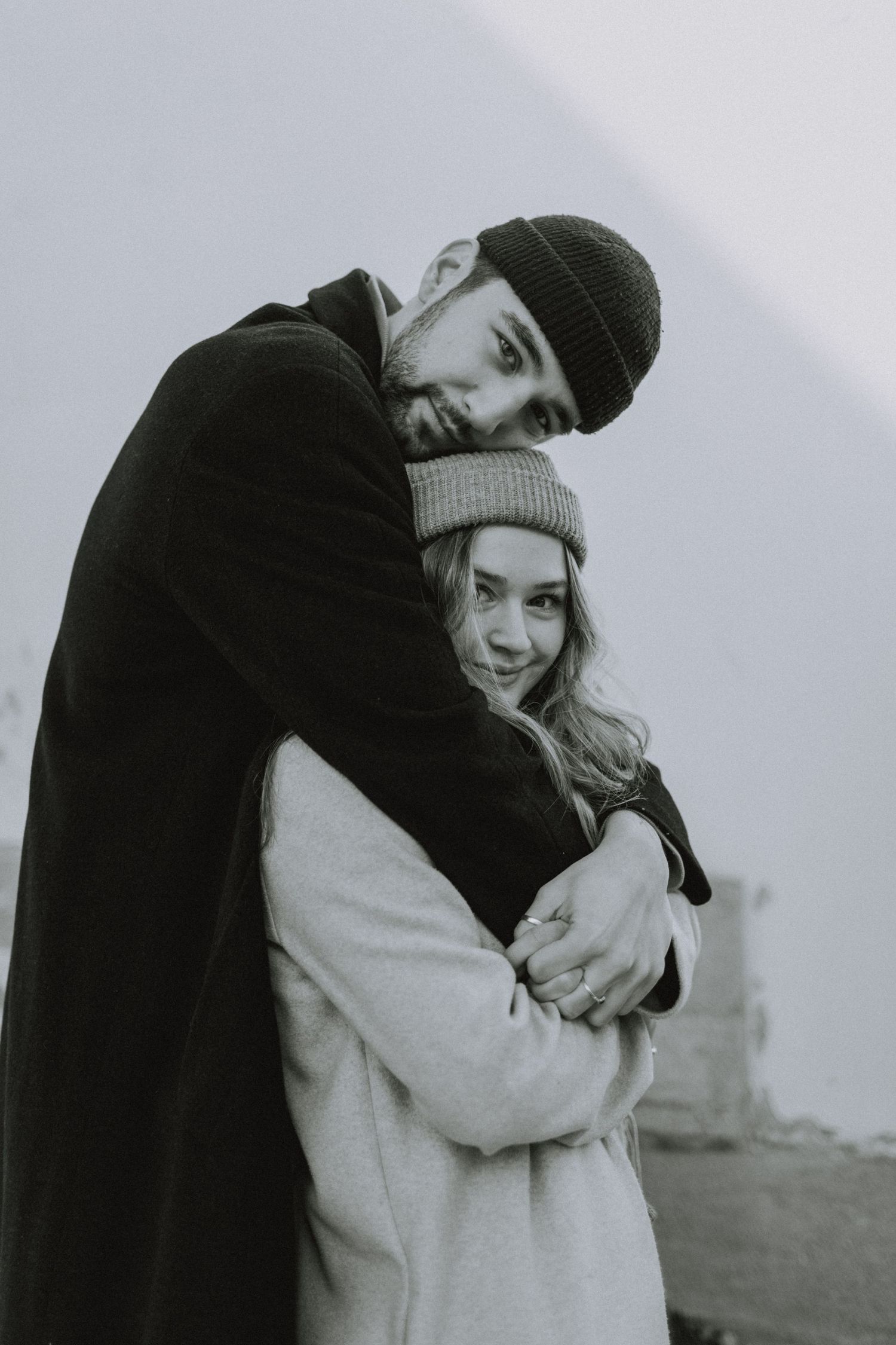 A romantic black and white portrait of a couple embracing outdoors while wearing winter coats and knit hats.