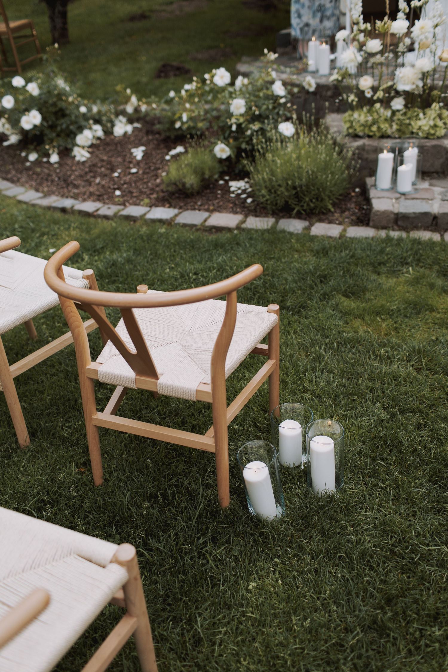 Wooden outdoor chair with cream cushions sits on green lawn next to white candles and blooming garden border.