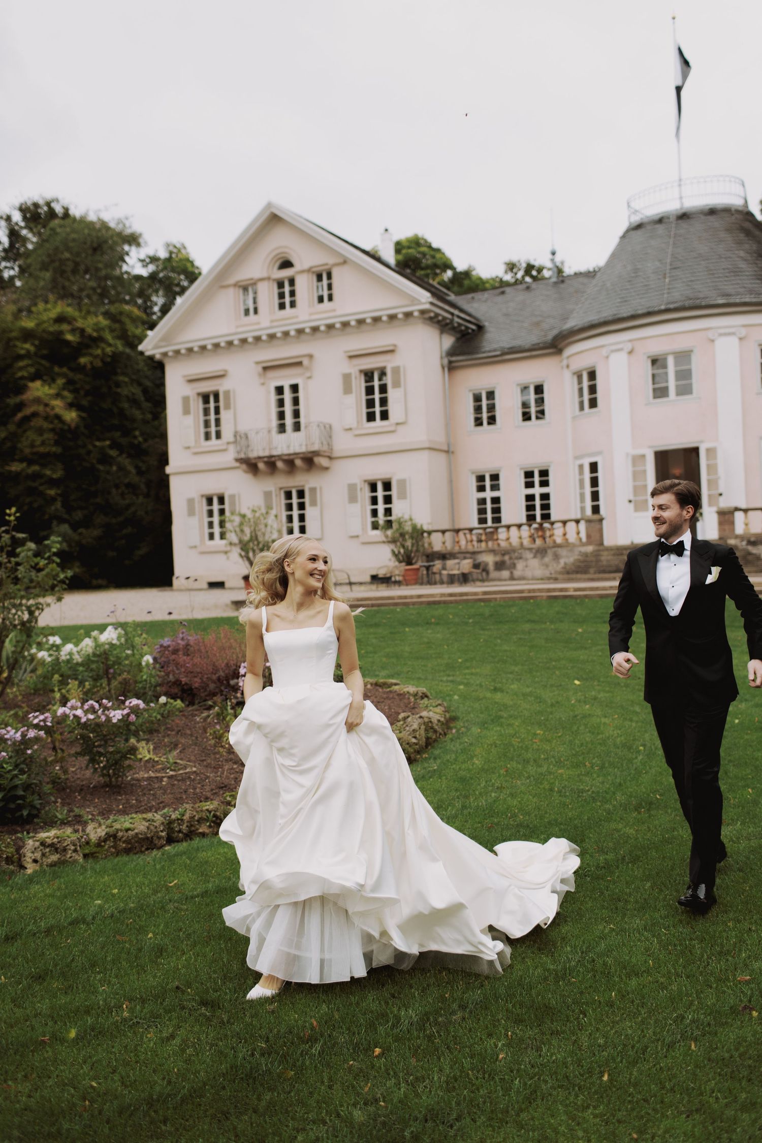 Couple walking on lawn in front of elegant white mansion during wedding celebration.