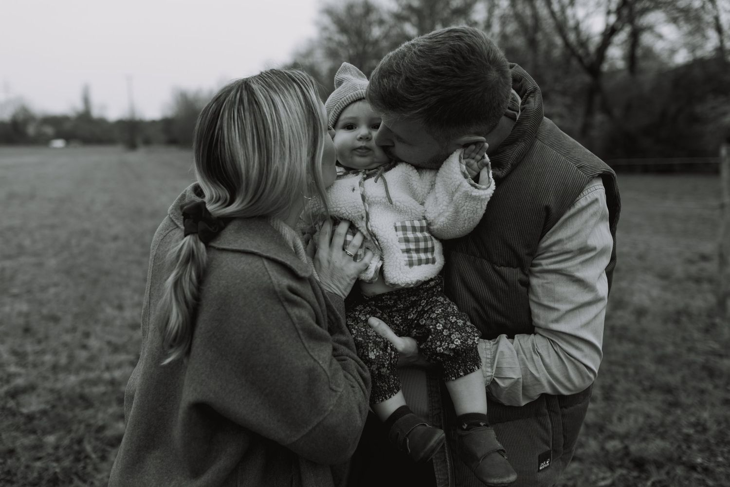 A tender black and white family moment captured outdoors in a field on an overcast day.