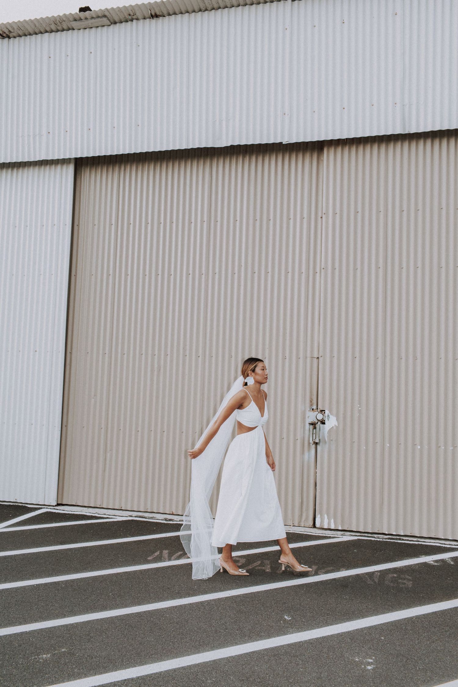 Bride in flowing white dress walking against industrial metal wall.