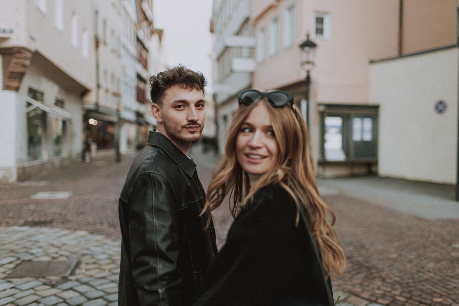 A couple in leather jackets smiling at the camera on a European cobblestone street.