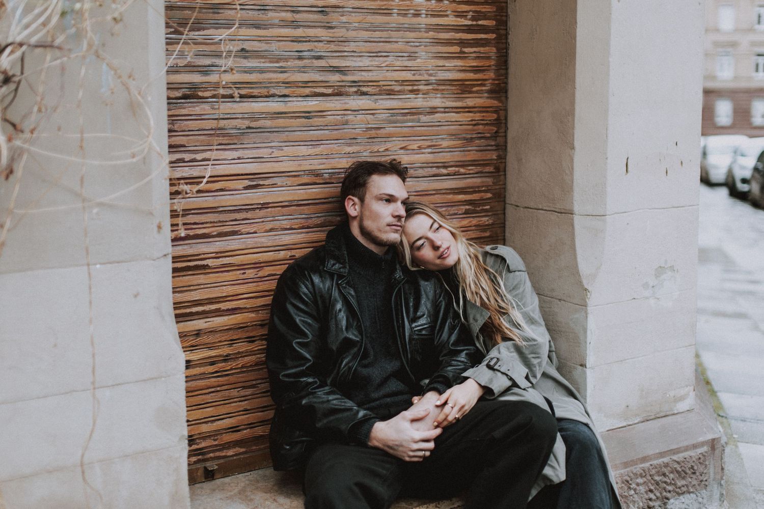 A couple in dark winter clothing sits intimately together against a wooden wall outdoors.