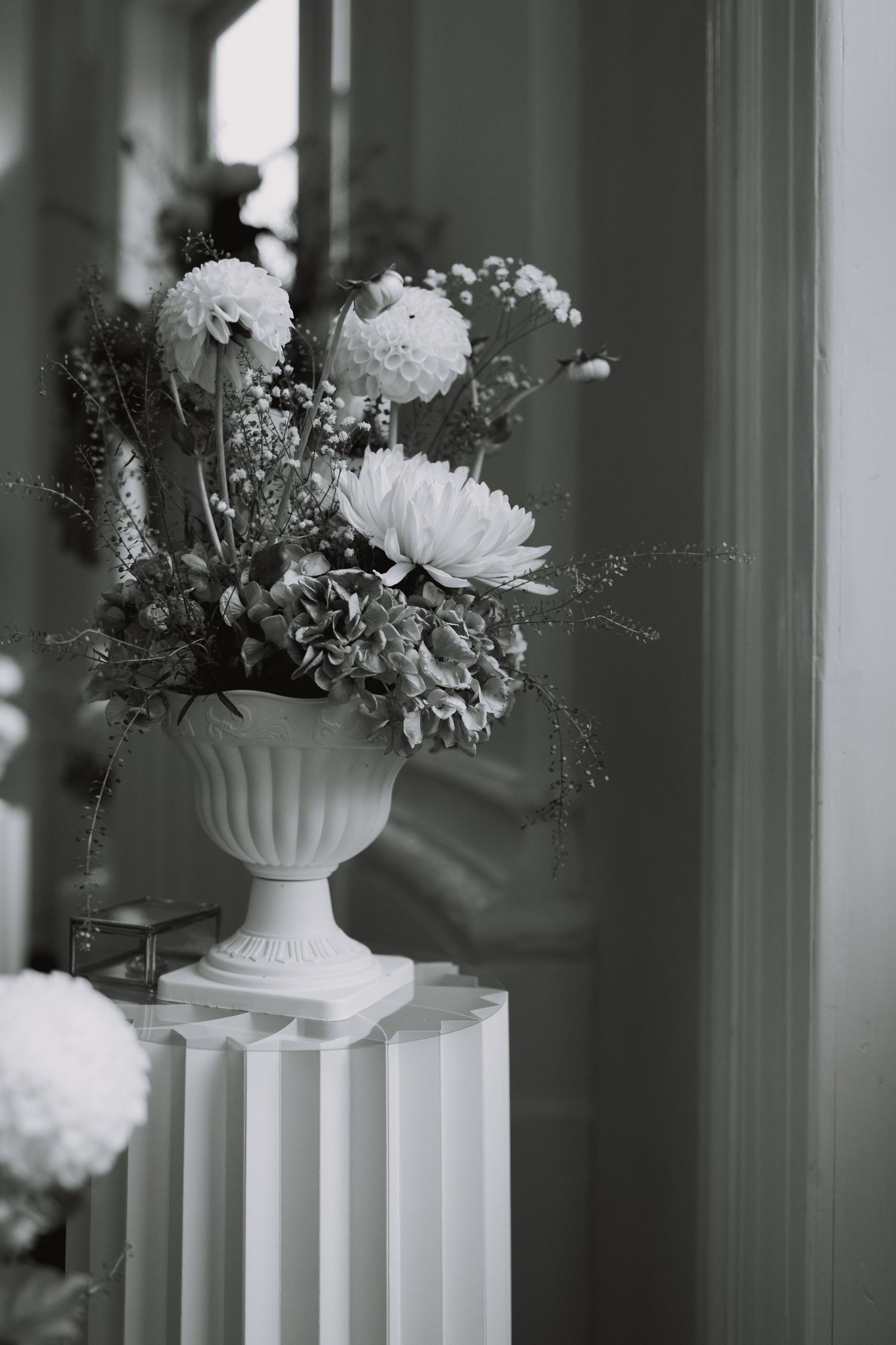 An elegant white floral arrangement in a classical urn vase against a window.