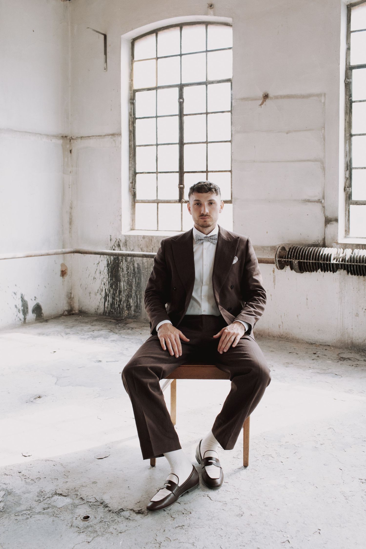 A groom in a dark suit sits on a wooden chair in an industrial white room.