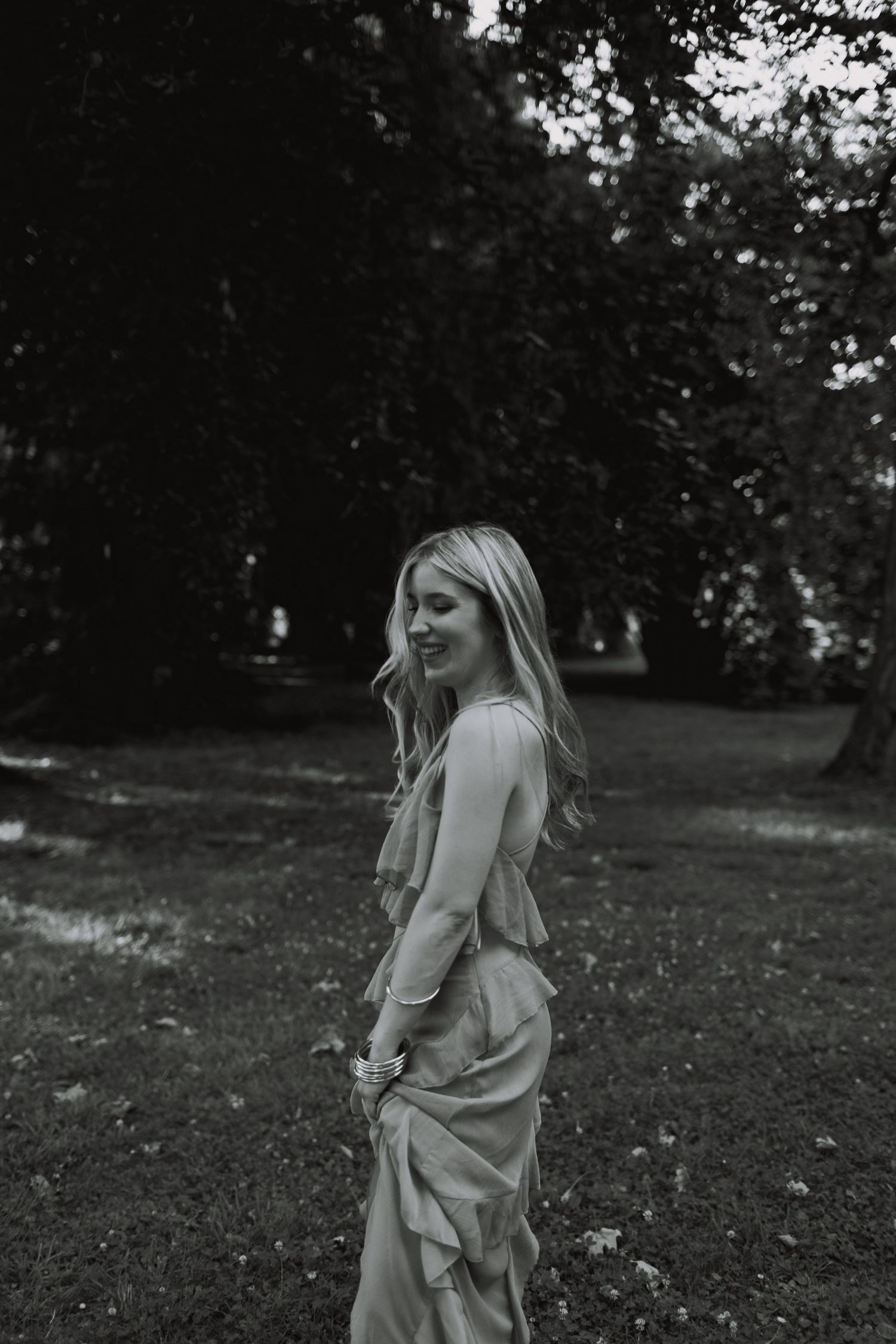 A person in a light-colored dress spins joyfully in a grassy outdoor setting with trees in the background.