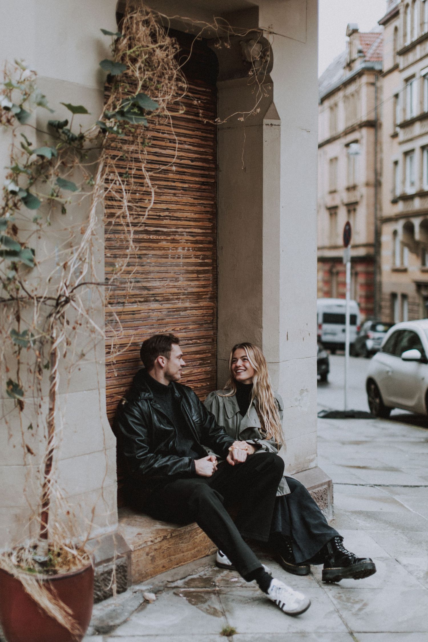 Two people in black coats sit and laugh together on stone steps against a brick wall in an urban setting.