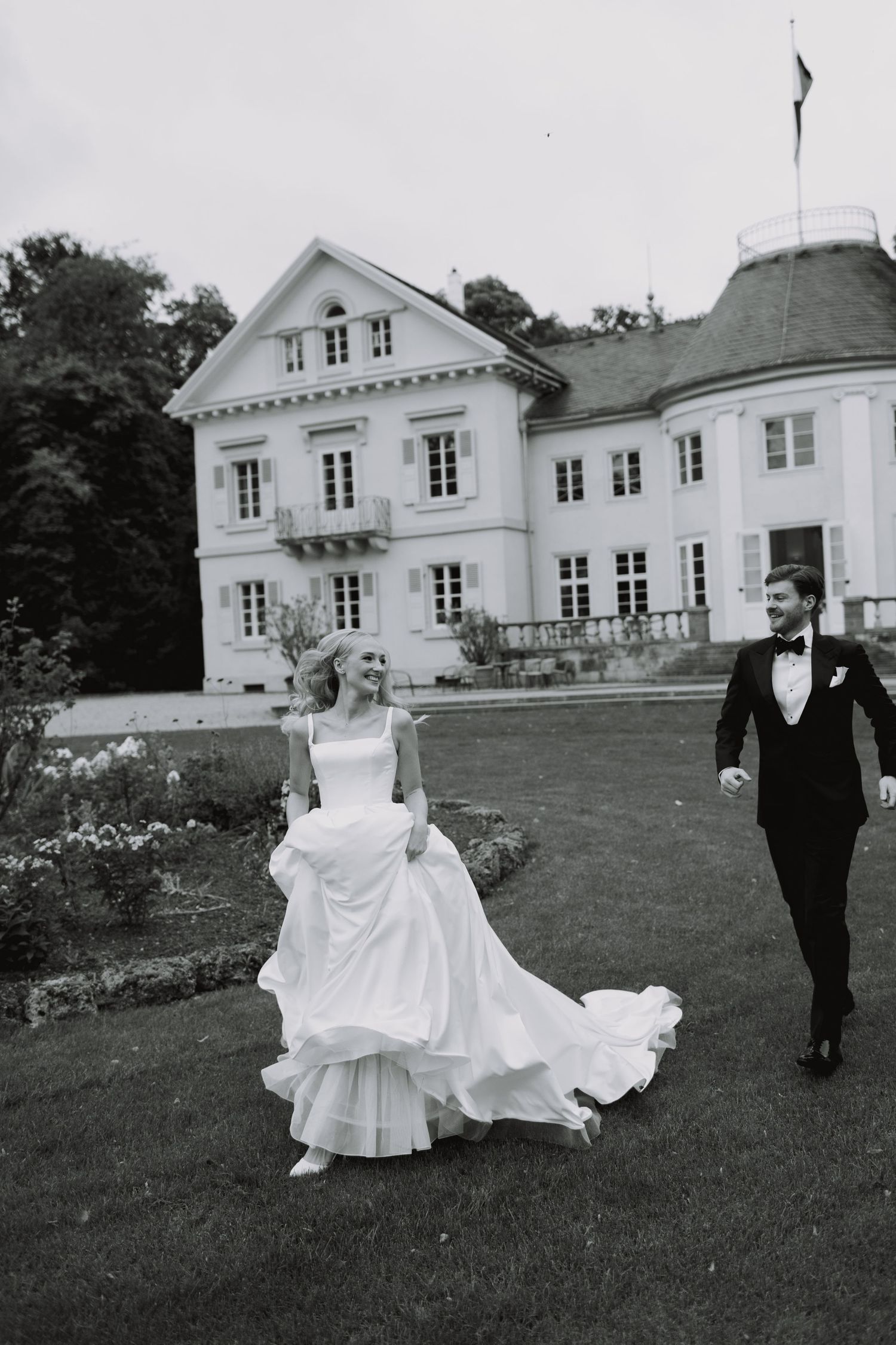 A couple walks across a grand manor house lawn in formal wedding attire.