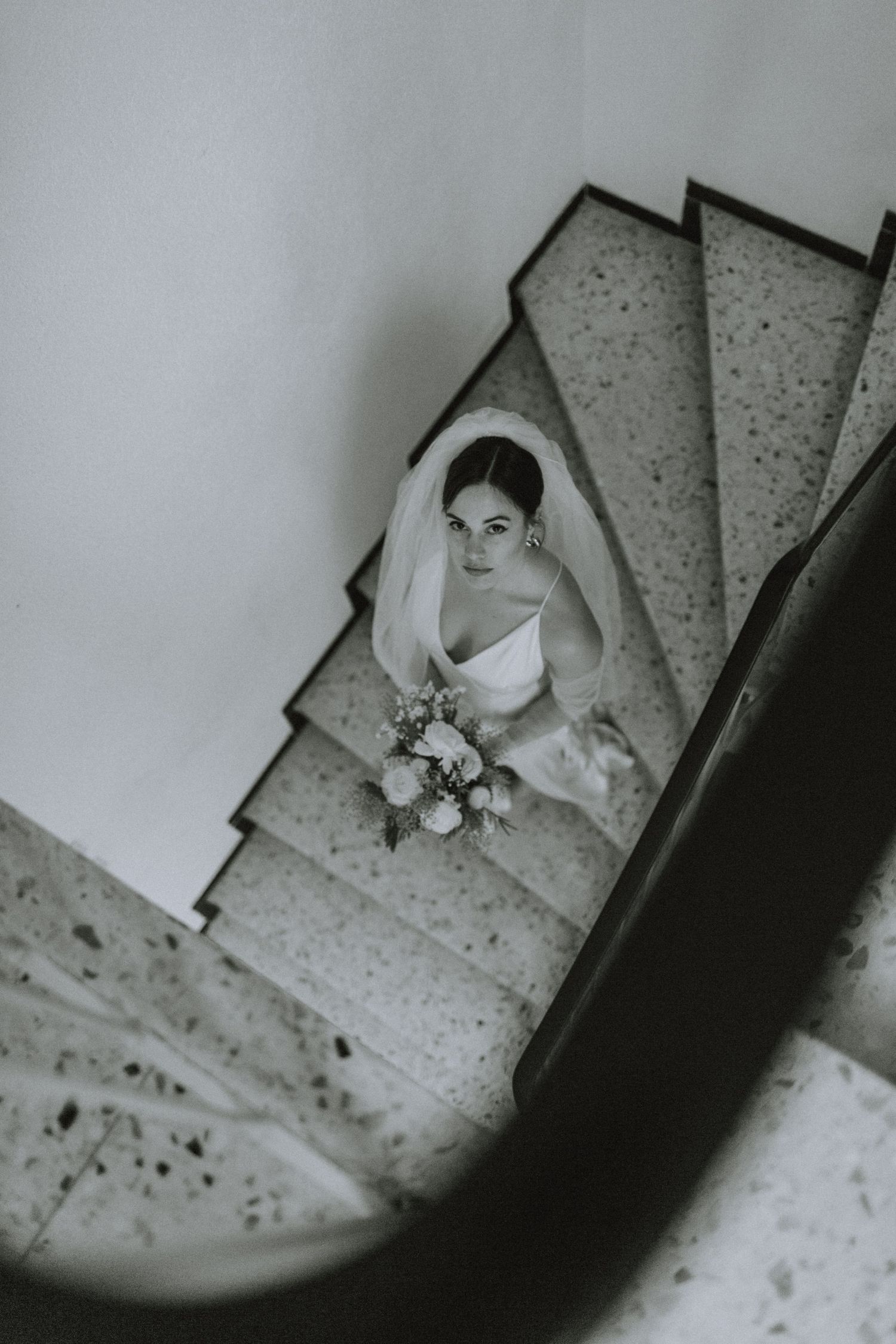 Black and white overhead view of someone in white dress descending spiral staircase.
