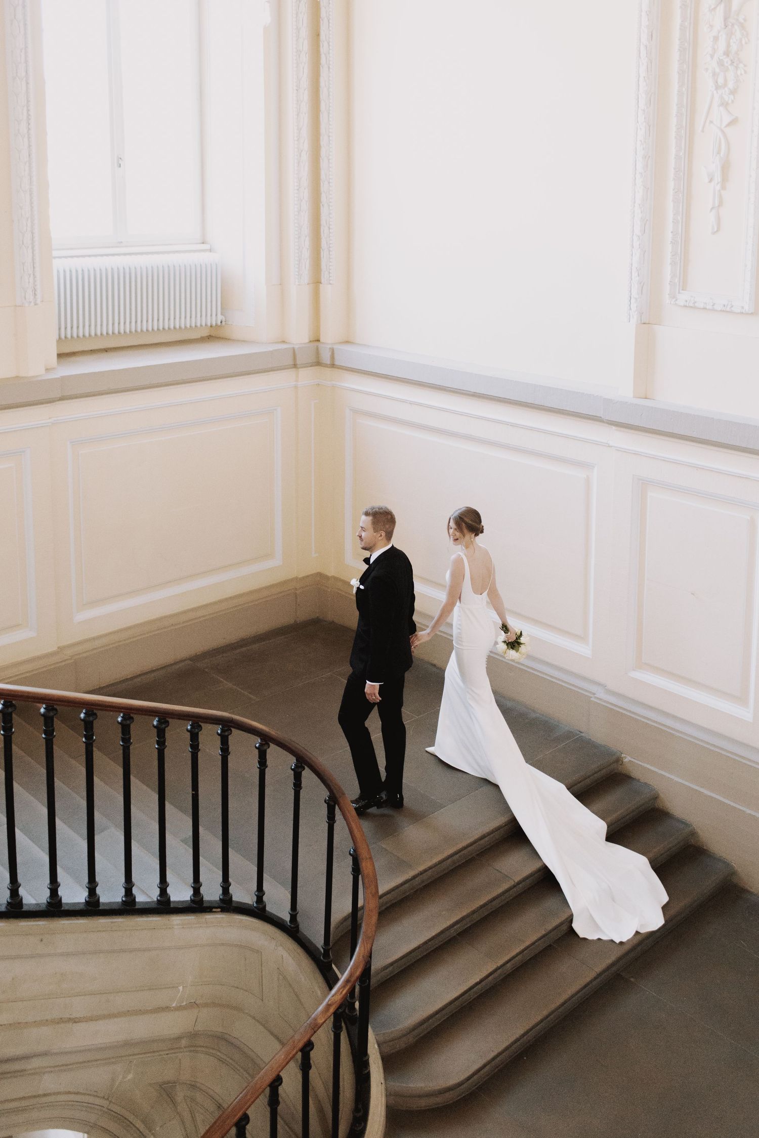 A couple in formal wedding attire walks up an elegant curved staircase with white walls and dark banister.
