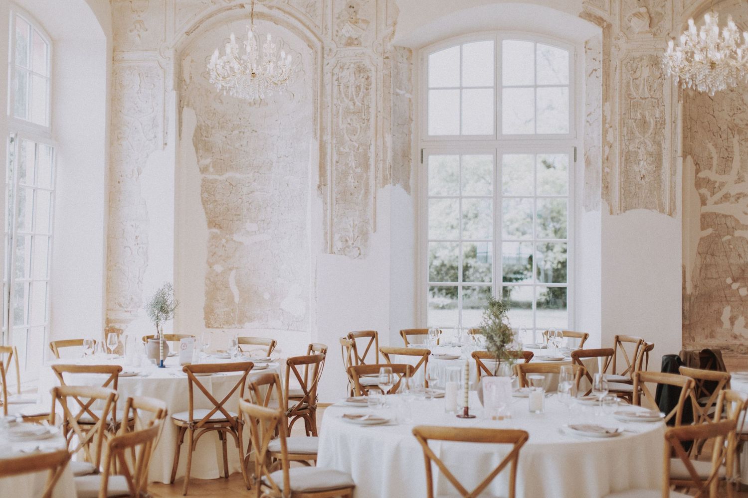 Elegant white wedding reception venue with crystal chandeliers, wooden cross-back chairs and large windows letting in natural light.