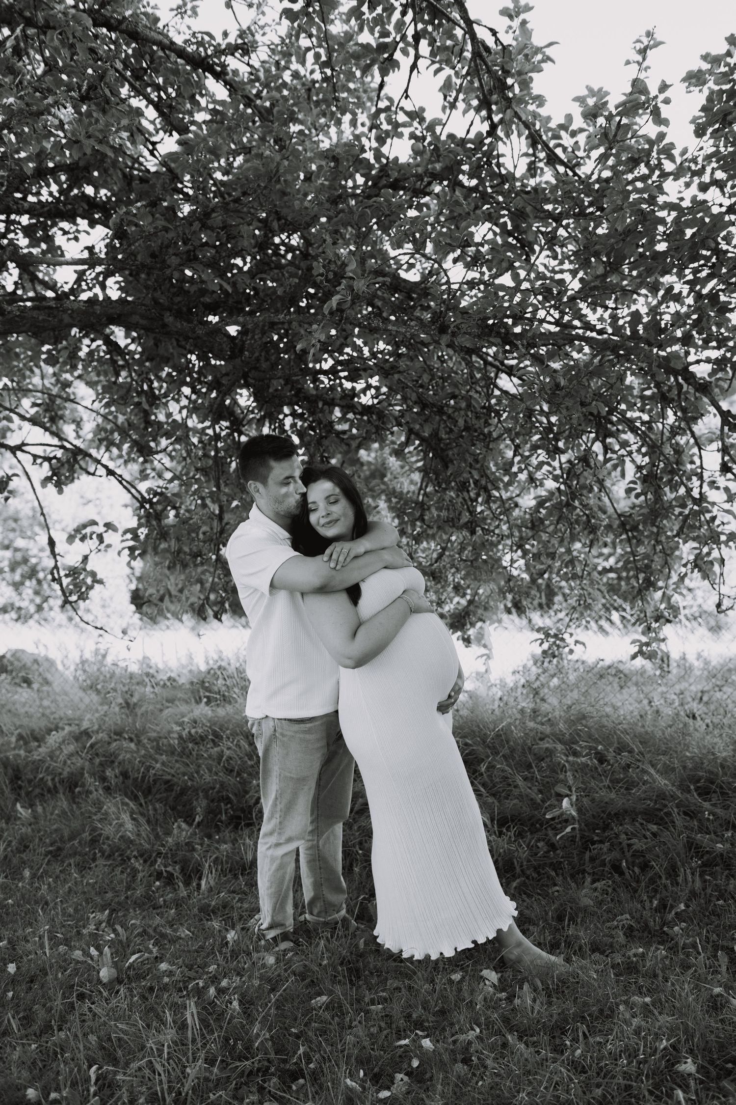 A romantic black and white maternity photo of a couple embracing under a tree in a natural outdoor setting.