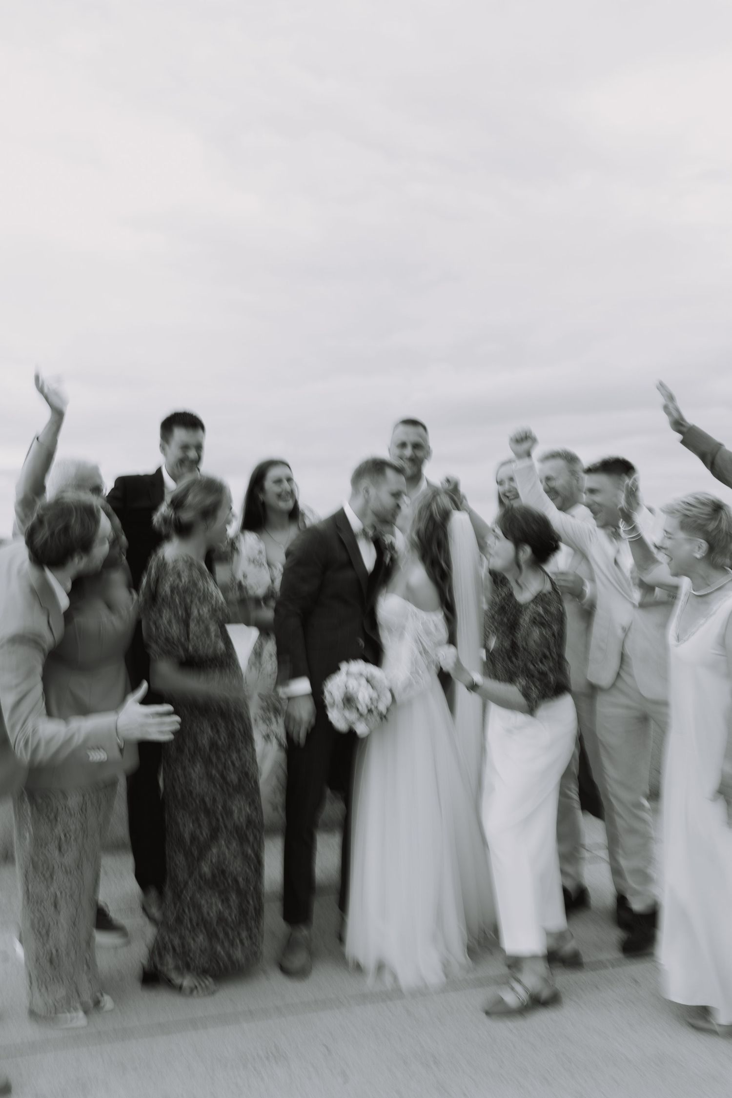 A wedding party poses in a line with their arms raised in celebration on a beach in black and white.