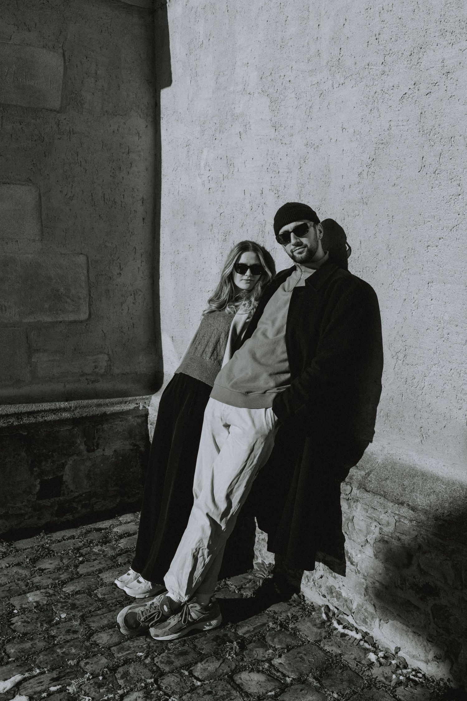 A stylish couple in winter coats posing against a white wall in an urban setting.