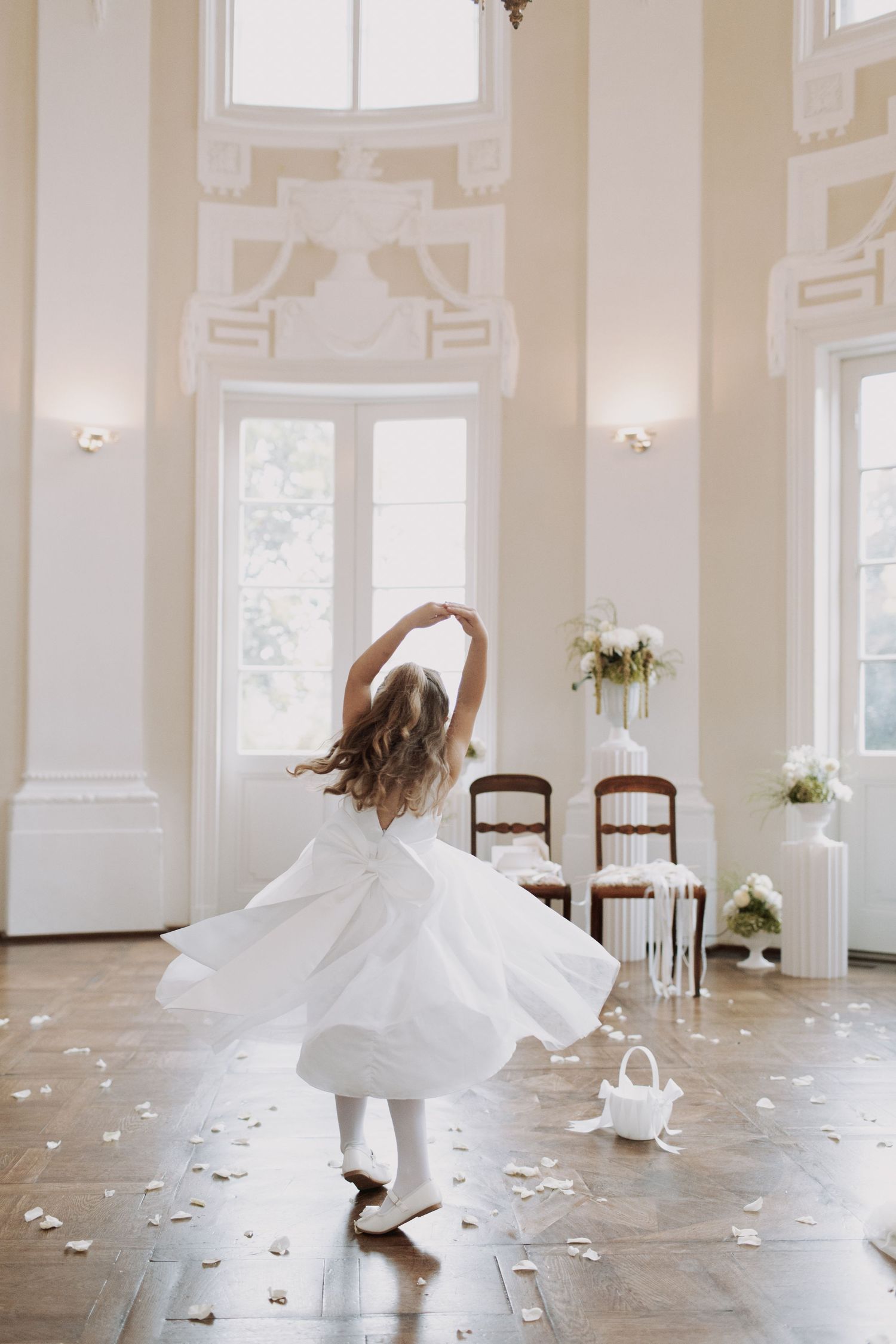 Joyful dancing figures in white dresses twirling in a bright, elegant room with scattered petals.