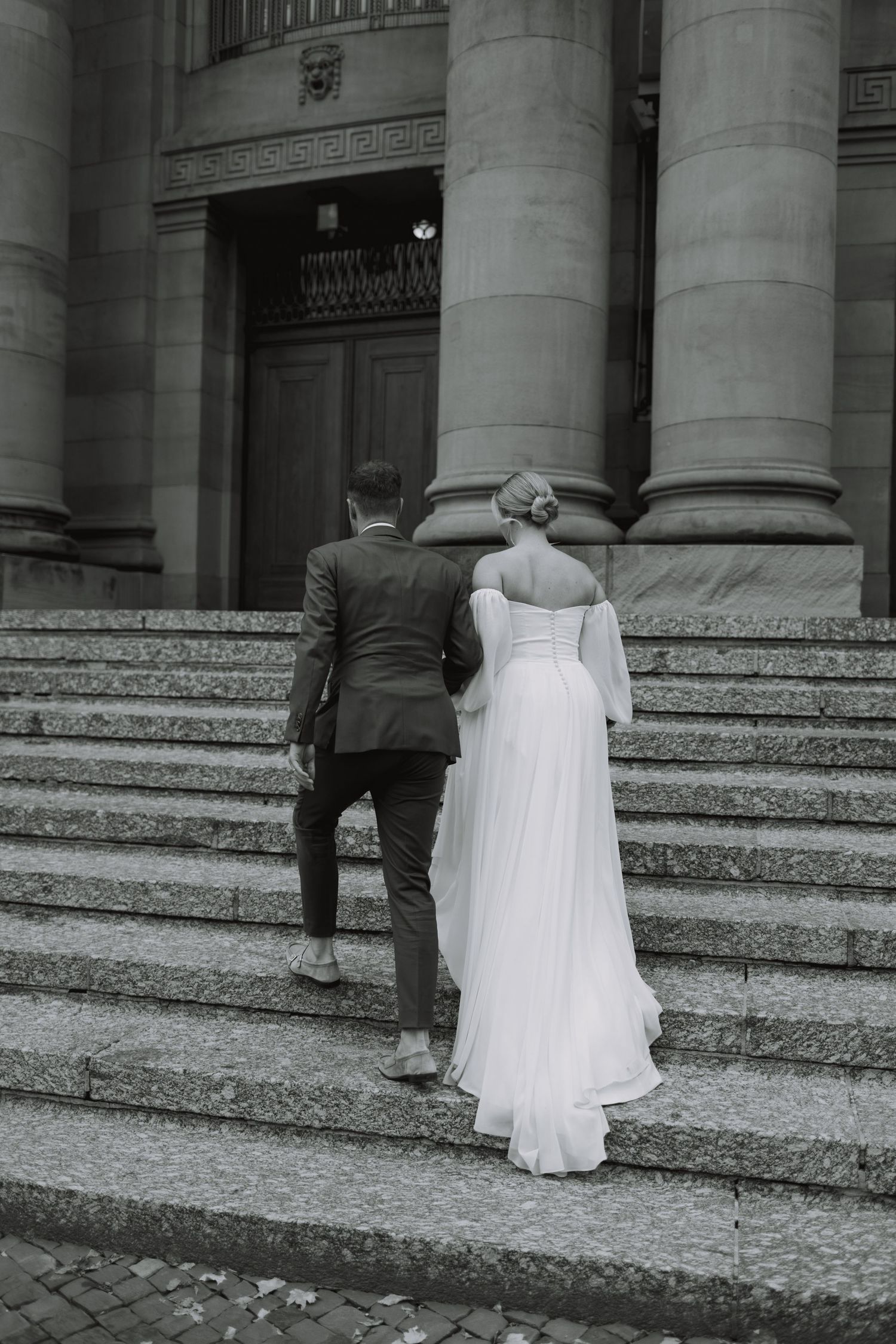 A couple ascends stone steps of a grand building in wedding attire.