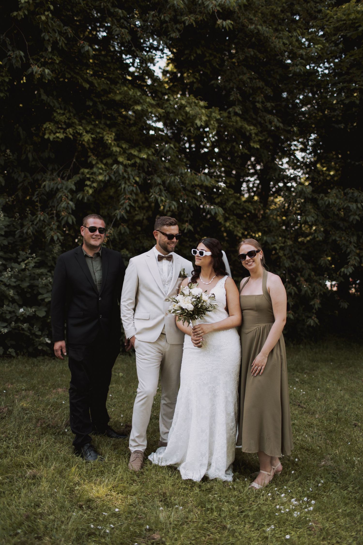 A group poses together outdoors in formal attire including white and neutral colored dresses against a wooded backdrop.
