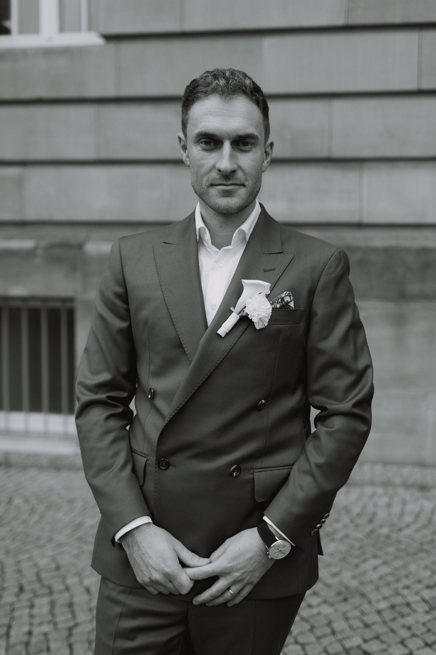 Groom in a dark suit with boutonniere poses against stone building.