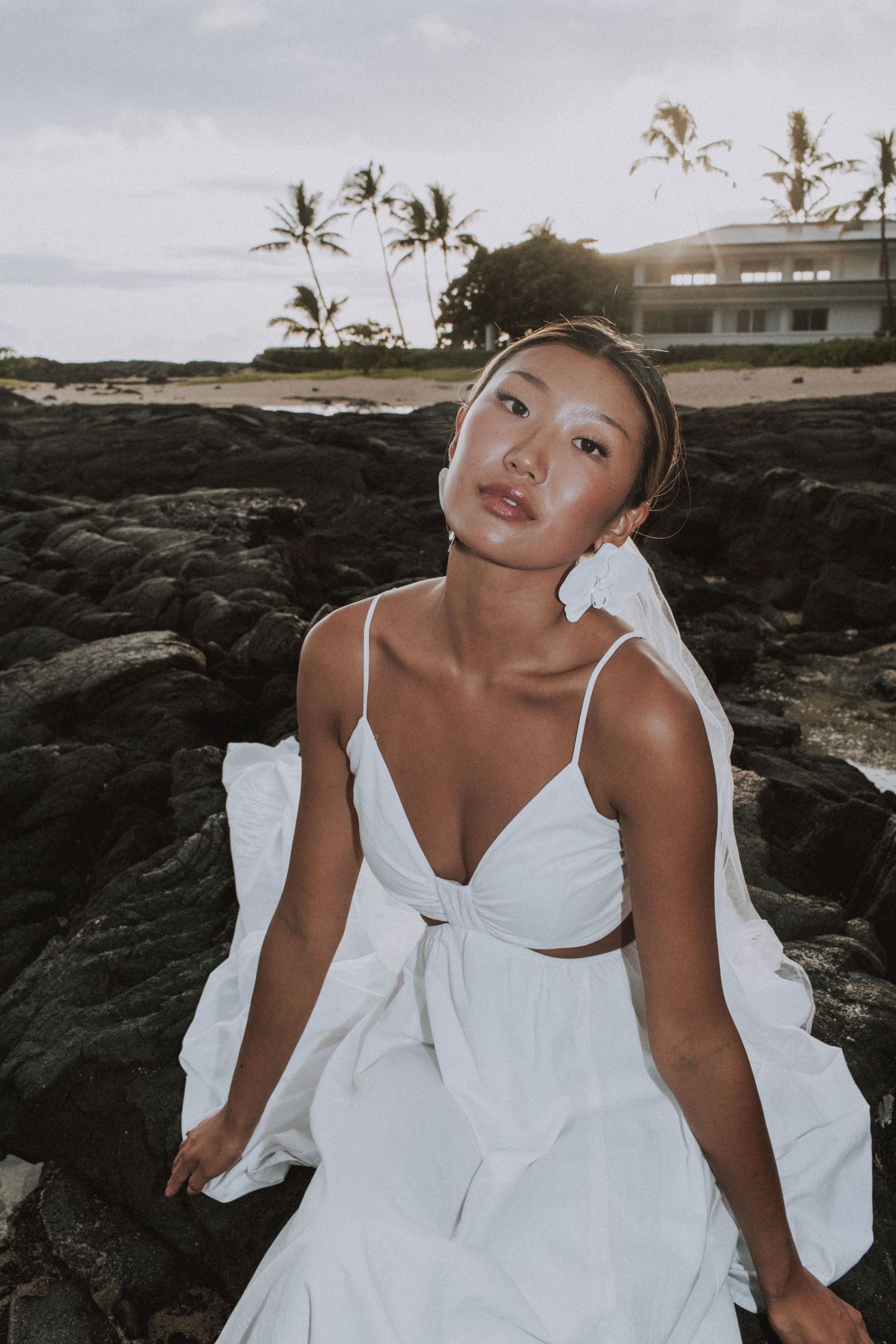 Bride in white dress poses dramatically on volcanic rocks near the ocean at sunset.