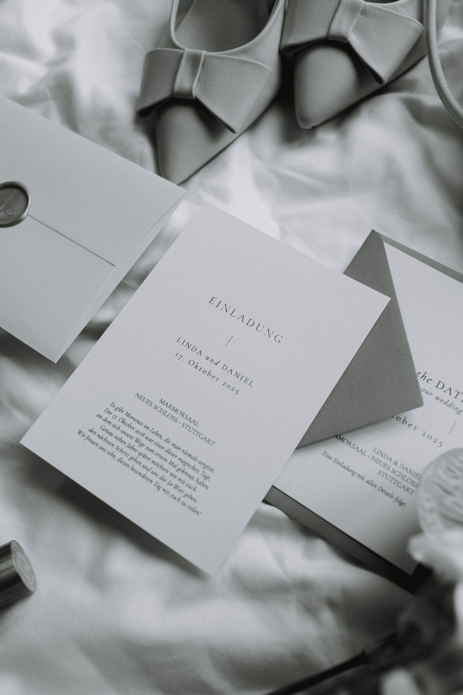 Elegant wedding invitation cards and gray bow heels arranged on white fabric.