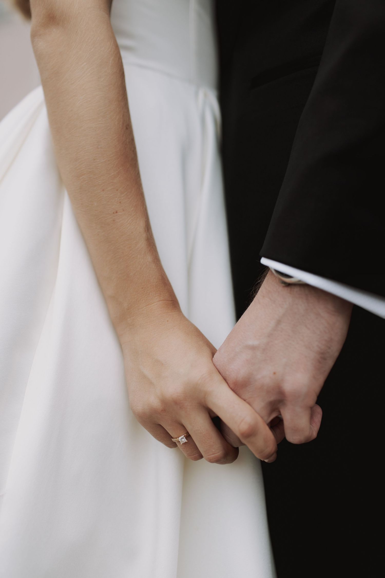 Close-up of newlyweds holding hands, showing wedding ring and formal attire details.
