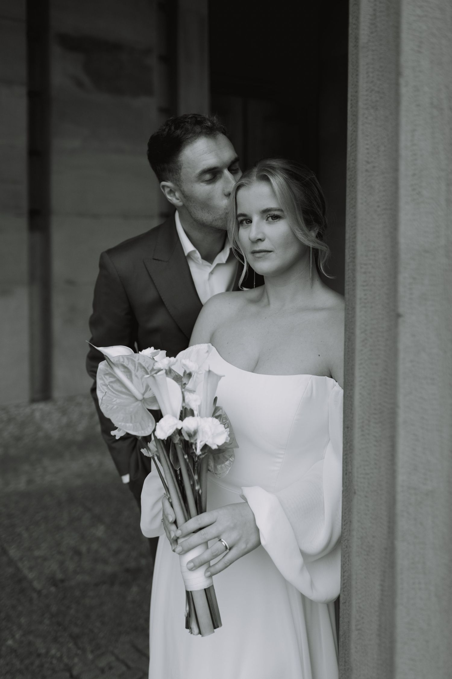 Black and white wedding portrait of couple embracing, bride in off-shoulder dress holding calla lily bouquet.