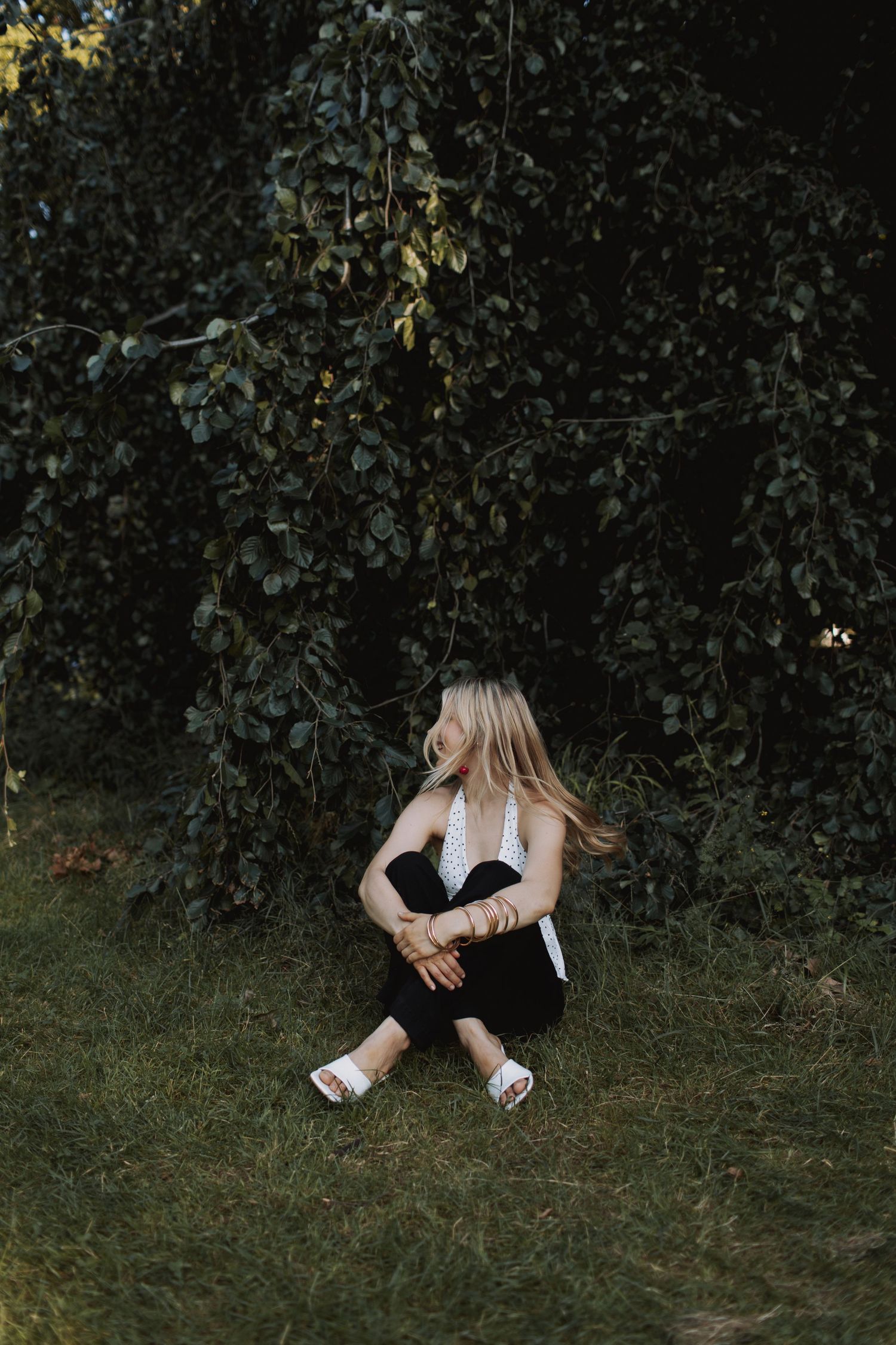 A person in dark clothing sits on grass against a shadowy background of trees and foliage.