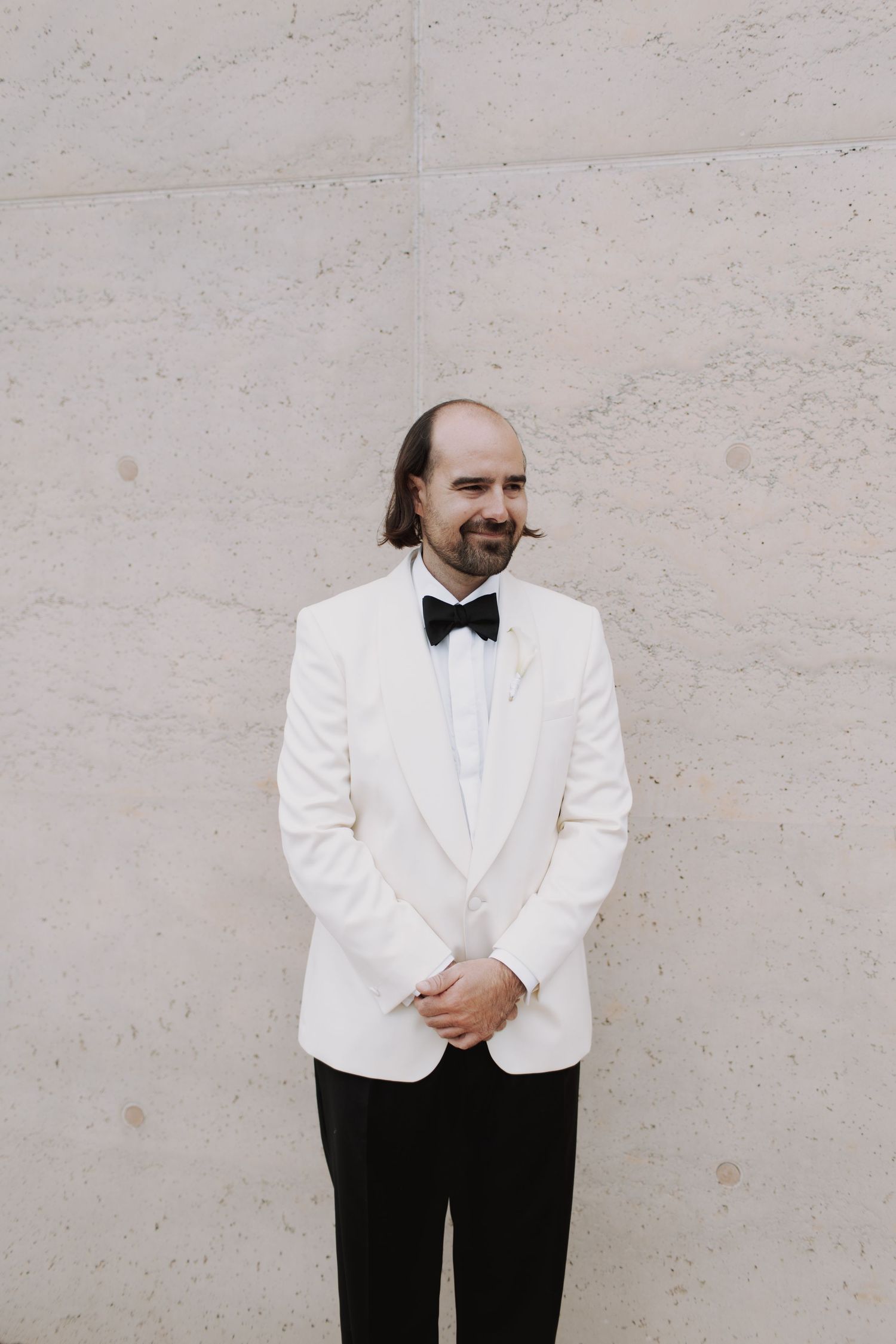 Groom in classic white dinner jacket and black bow tie poses against light colored wall.
