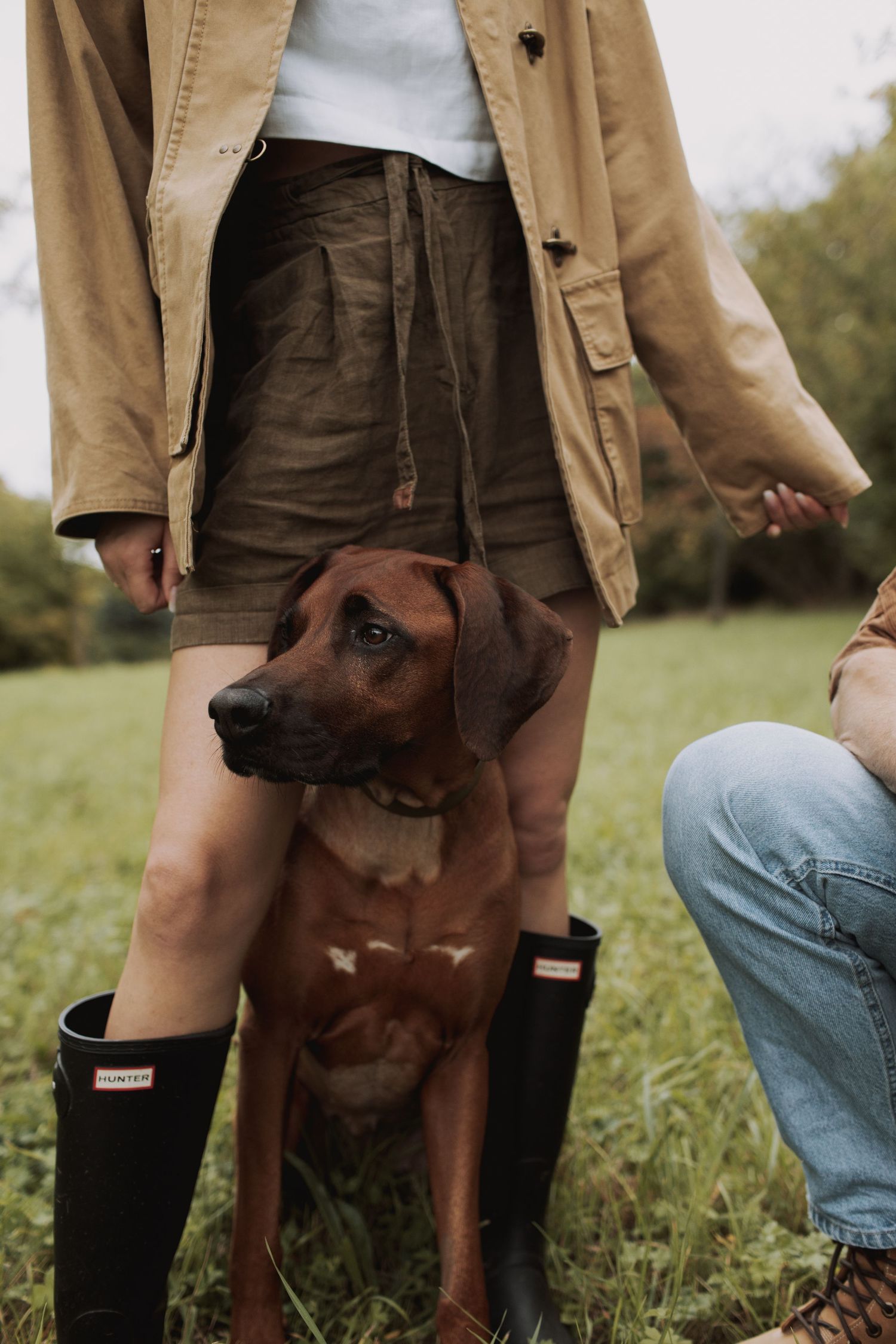 A brown dog stands between two people wearing rain boots and casual attire in a grassy outdoor setting.