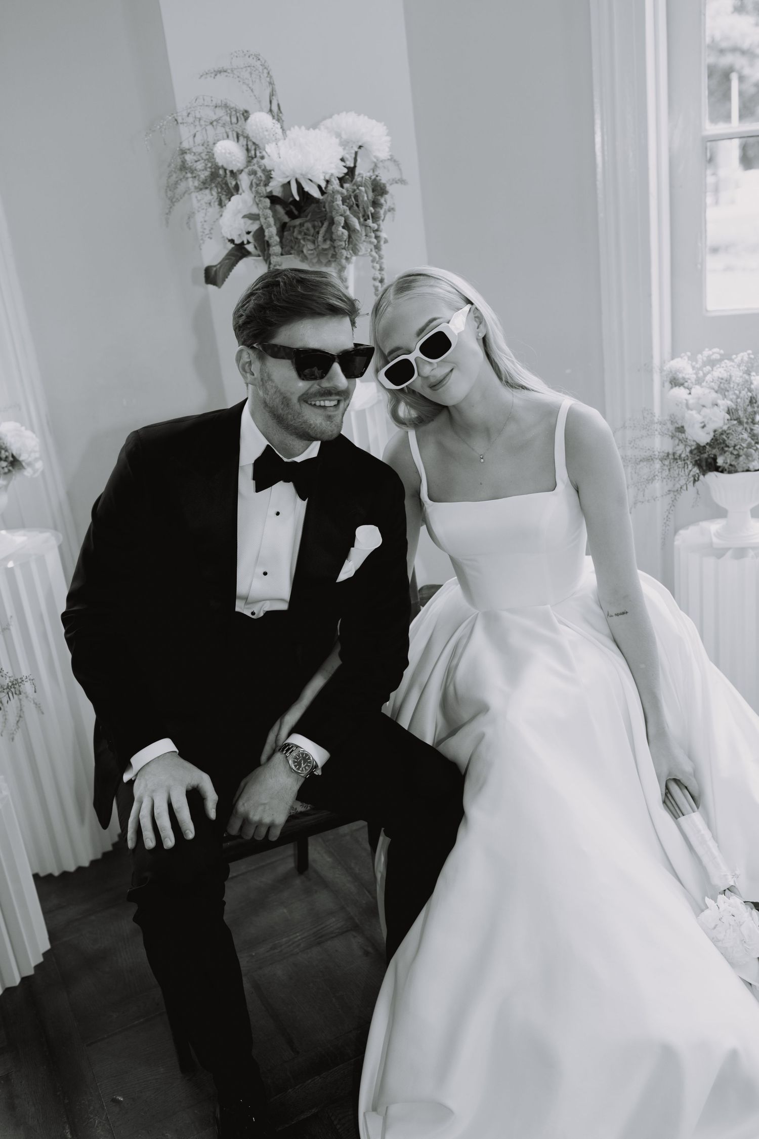 A stylish couple wearing sunglasses poses in formal wedding attire.
