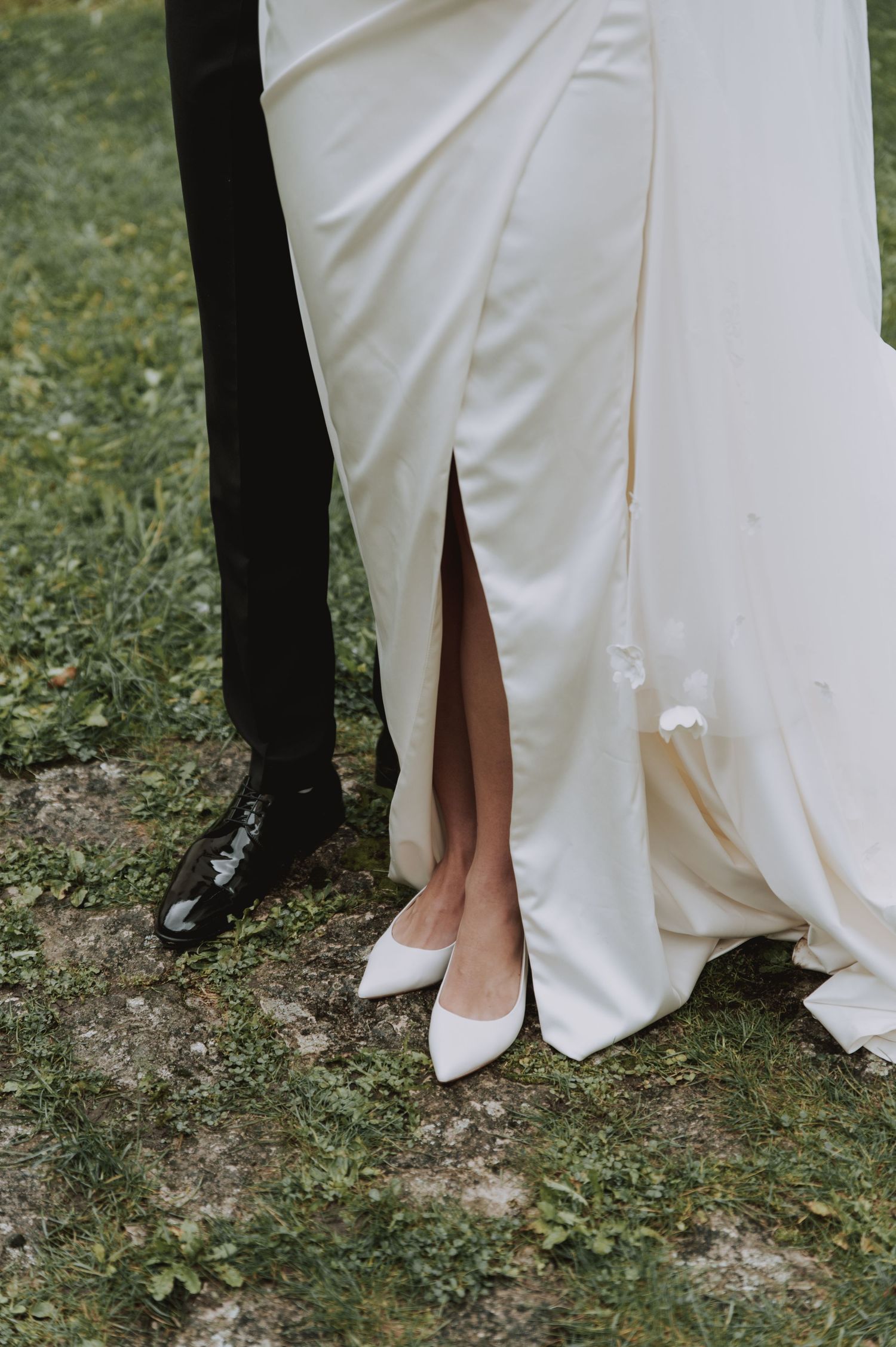 Close-up of white wedding dress slit and shoes next to black formal shoes on grass.