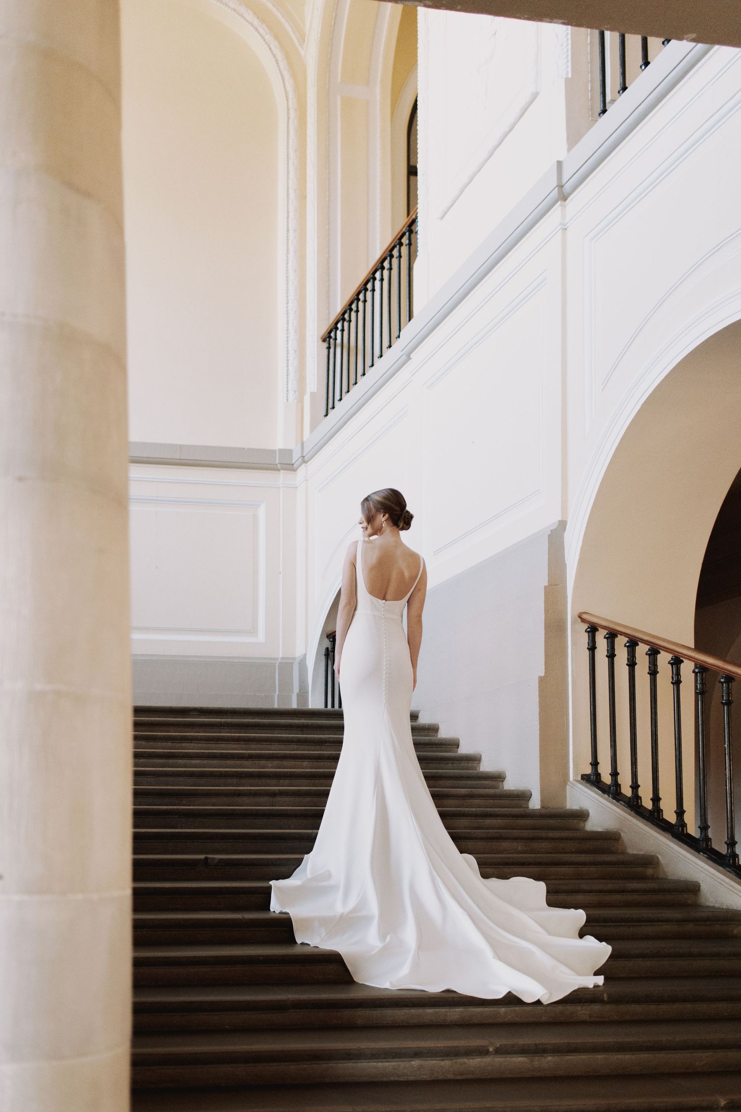 Elegant bride in flowing white gown stands gracefully on dark stairs against white architectural columns.