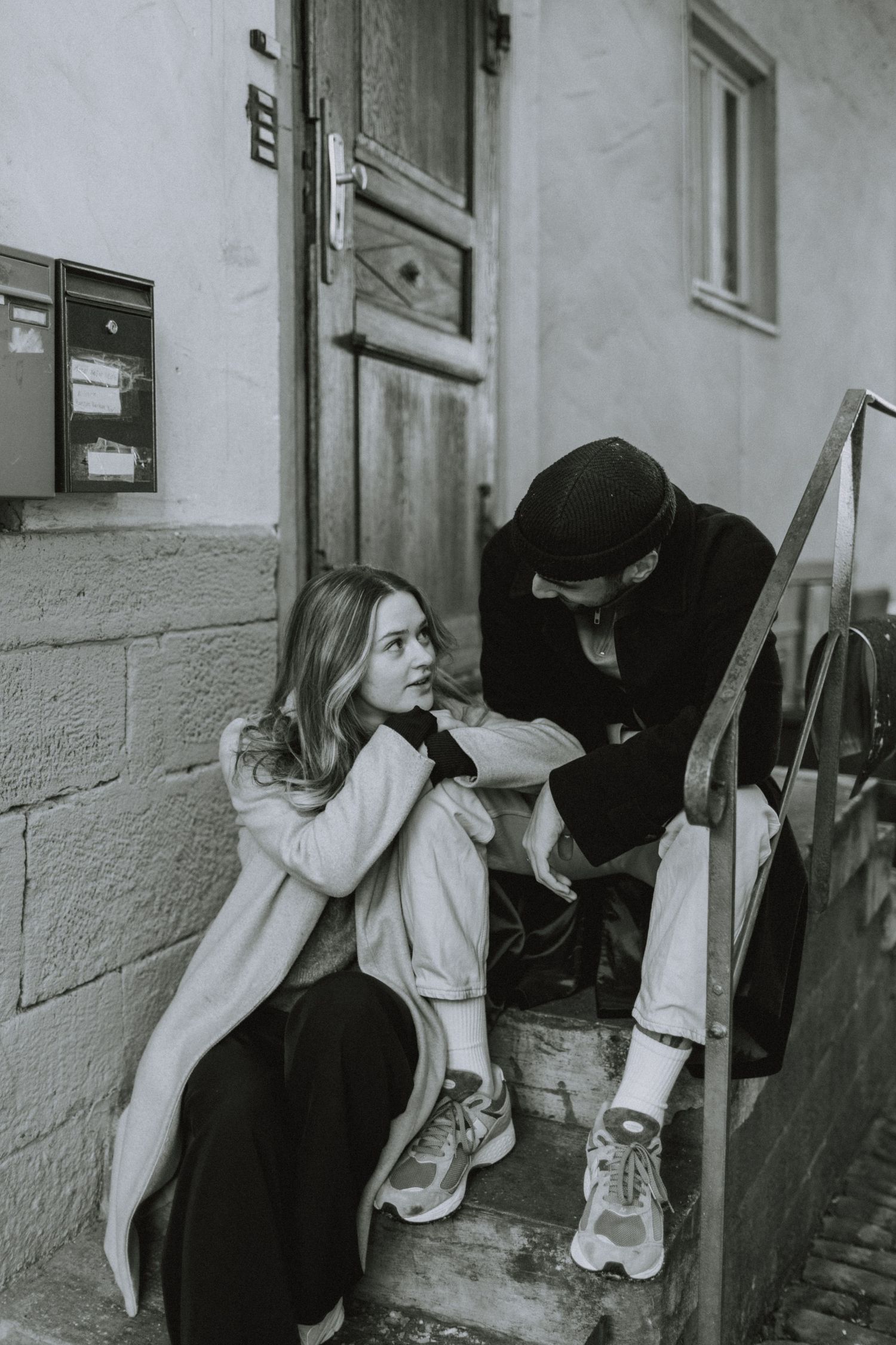 A candid black and white photo of a couple sitting on outdoor steps in casual winter attire.
