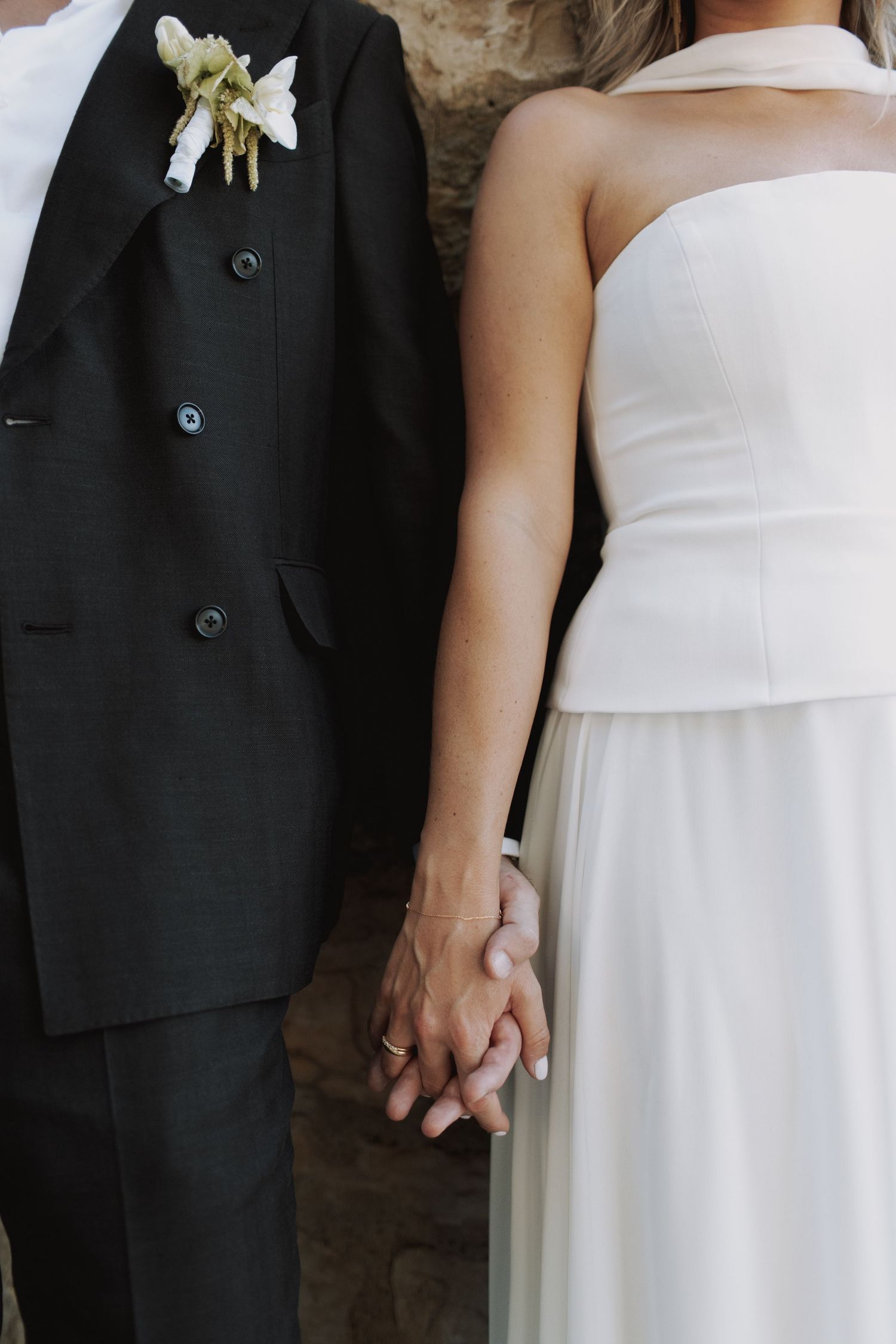 Close-up of newlyweds holding hands, showing black tuxedo with white boutonniere and white strapless wedding dress.