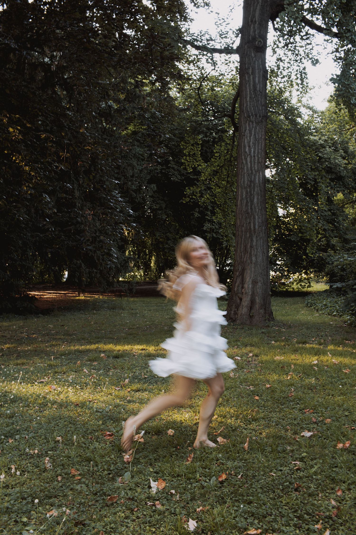 Motion blur captures a figure in a white dress twirling through a sunlit garden with tall trees and scattered leaves.