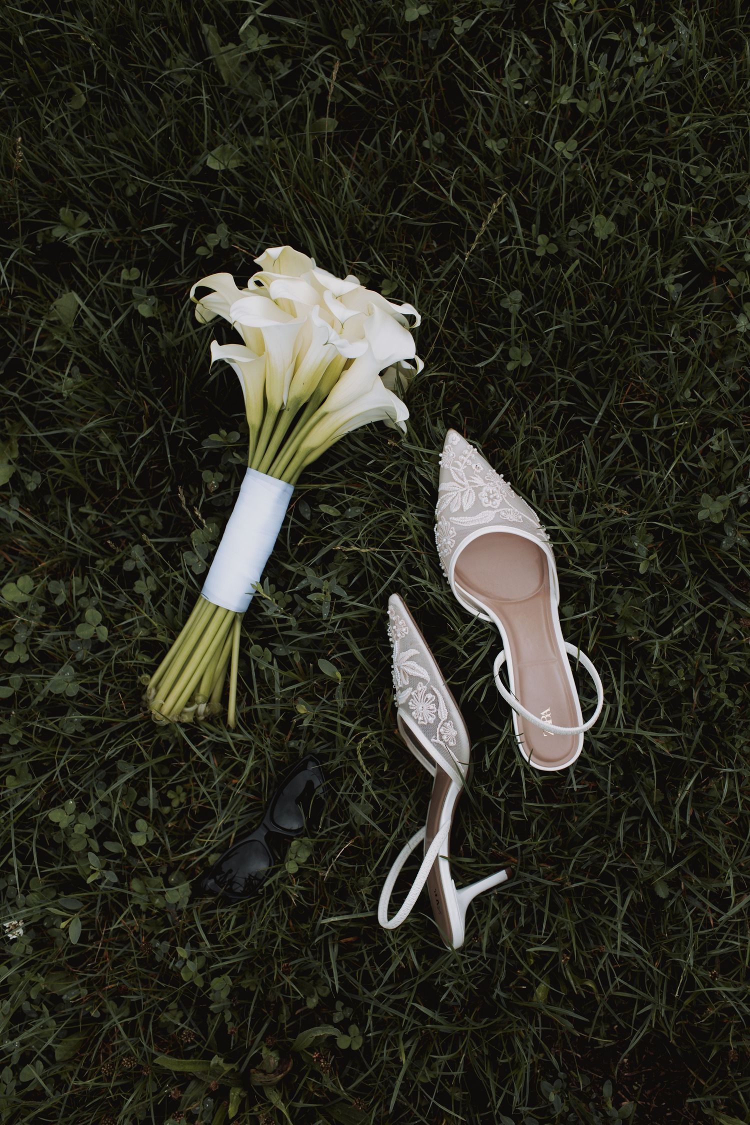 White bridal shoes and bouquet of calla lilies arranged on grass.