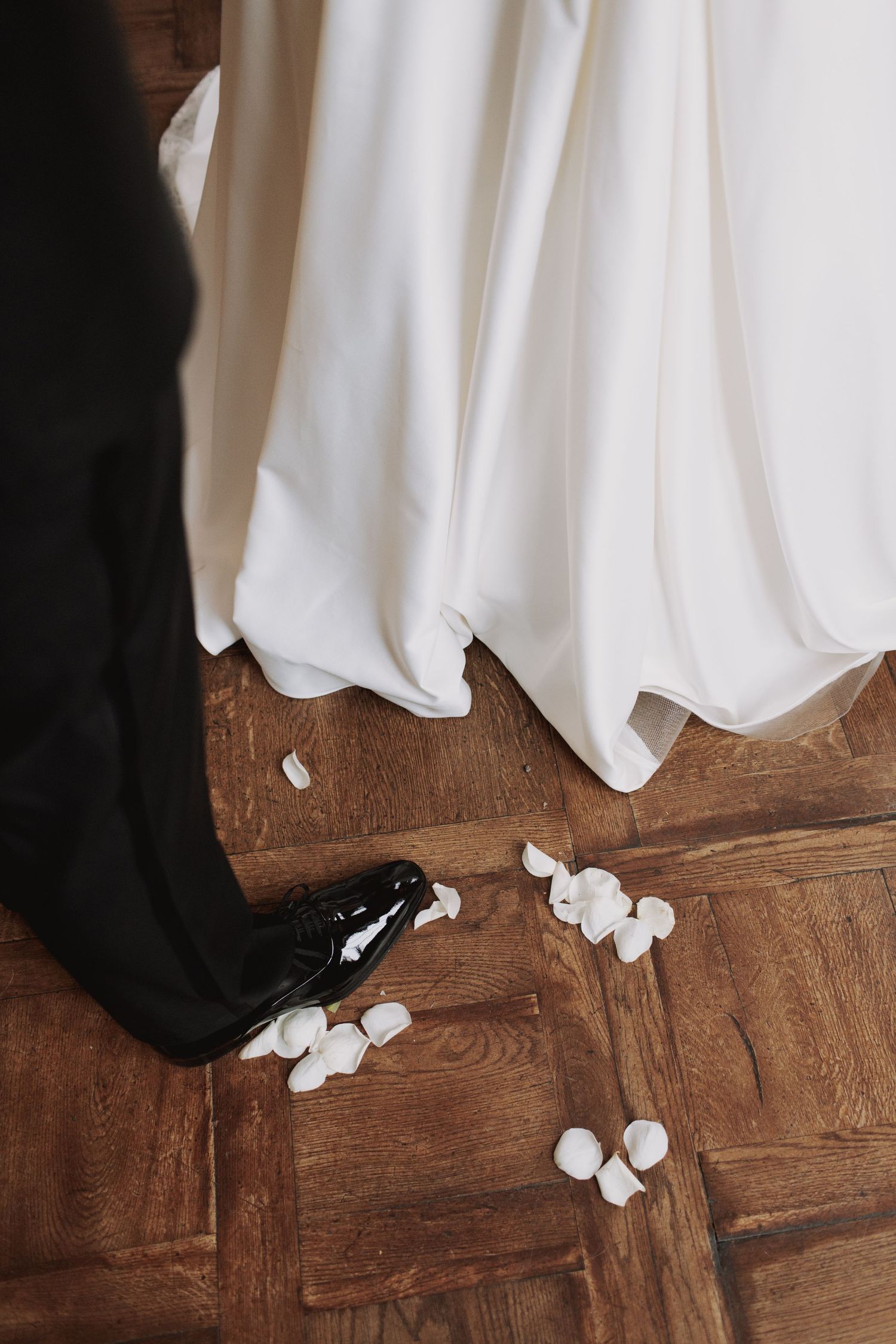White wedding dress and black shoes visible on a wooden floor with scattered rose petals.
