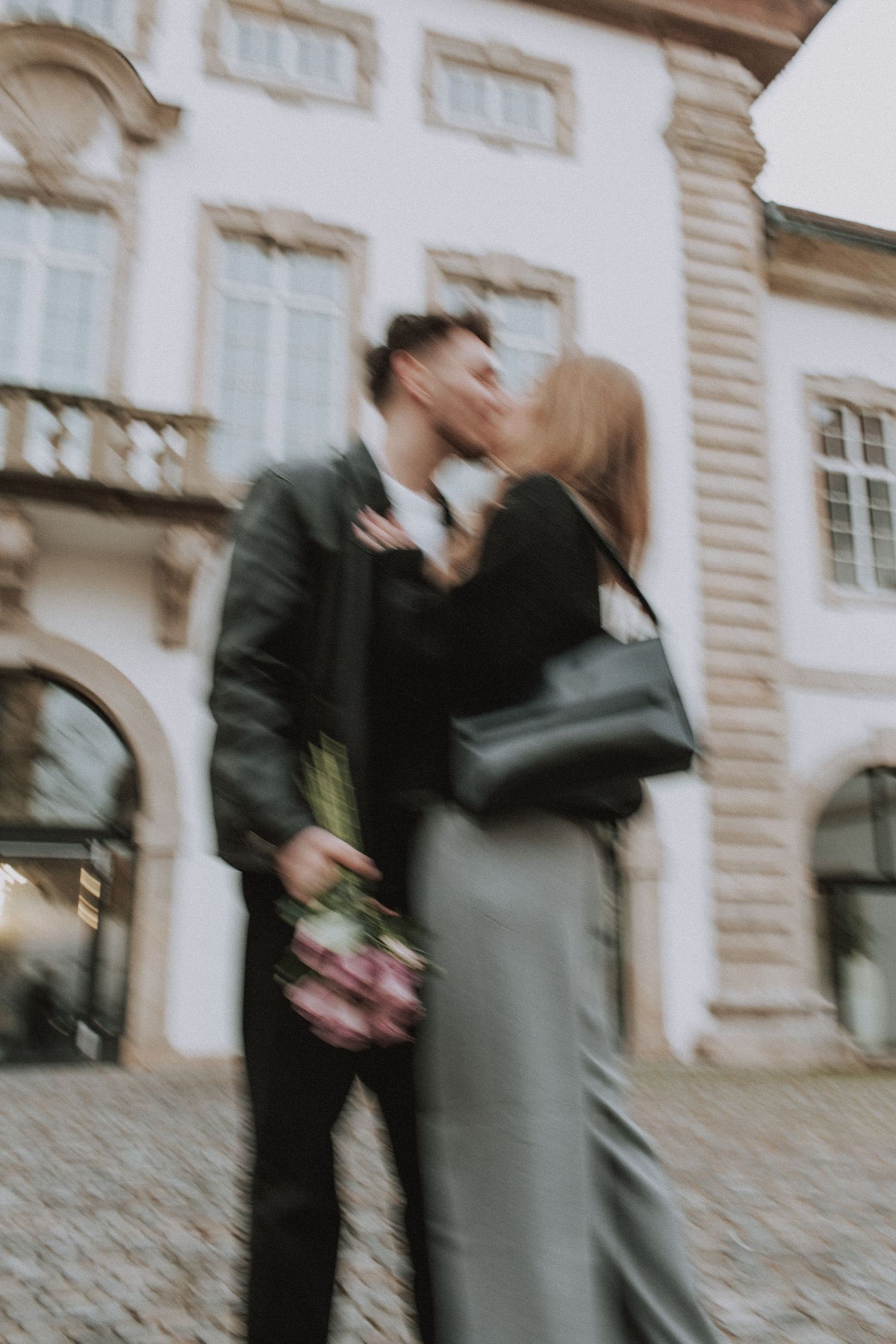 A romantic couple sharing a kiss while holding flowers outside a historic building.