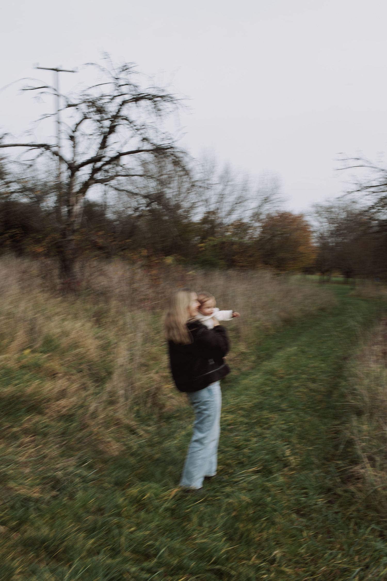 A person in a light colored jacket stands in a grassy field with bare trees in the background on an autumn day.