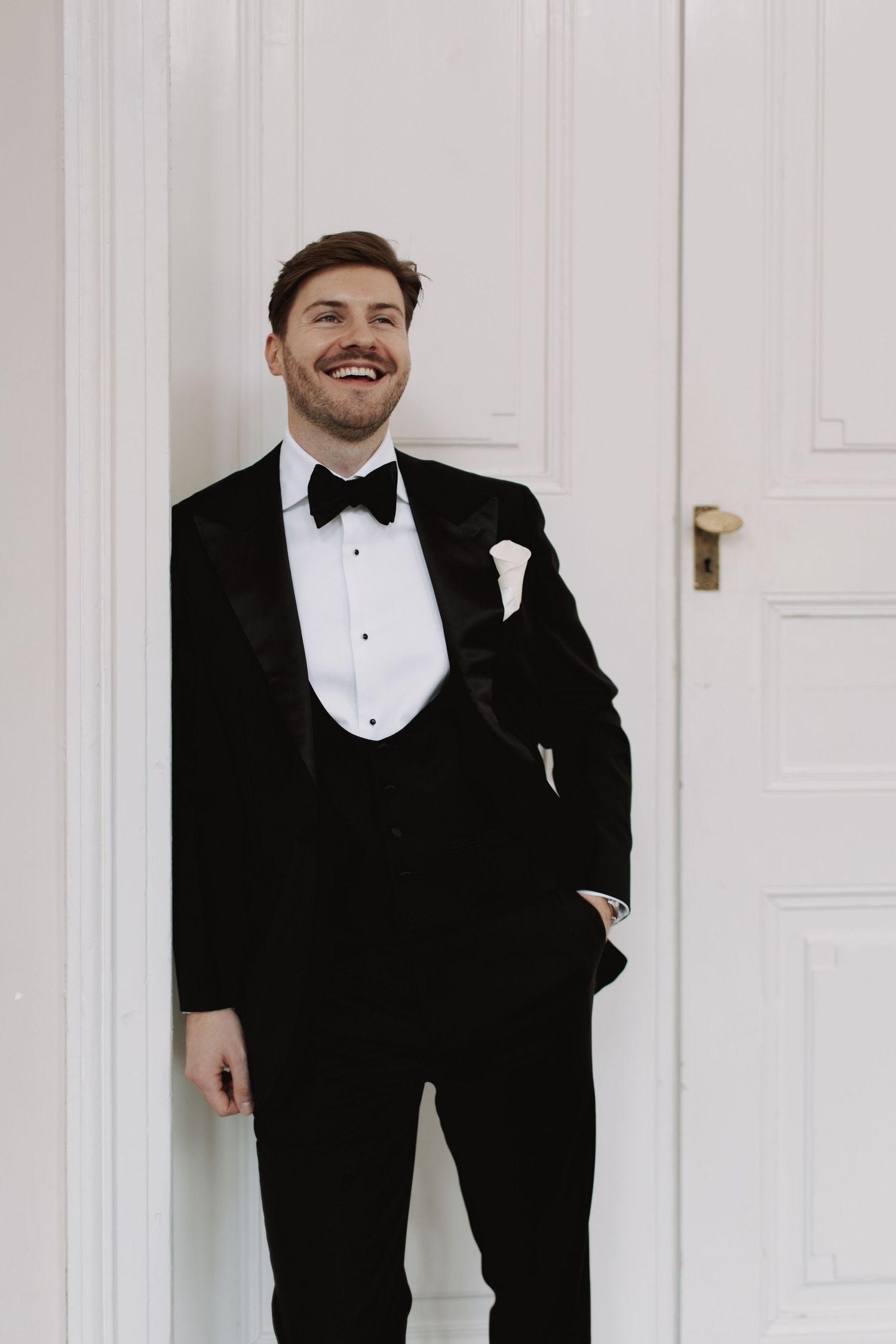 A formal portrait of a groom in classic black tuxedo against a white door.