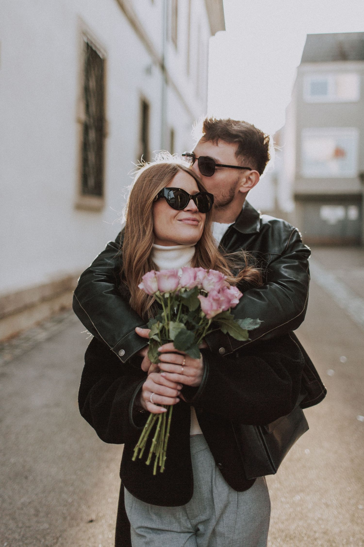 A couple in leather jackets sharing a tender moment while holding pink flowers on an urban street.
