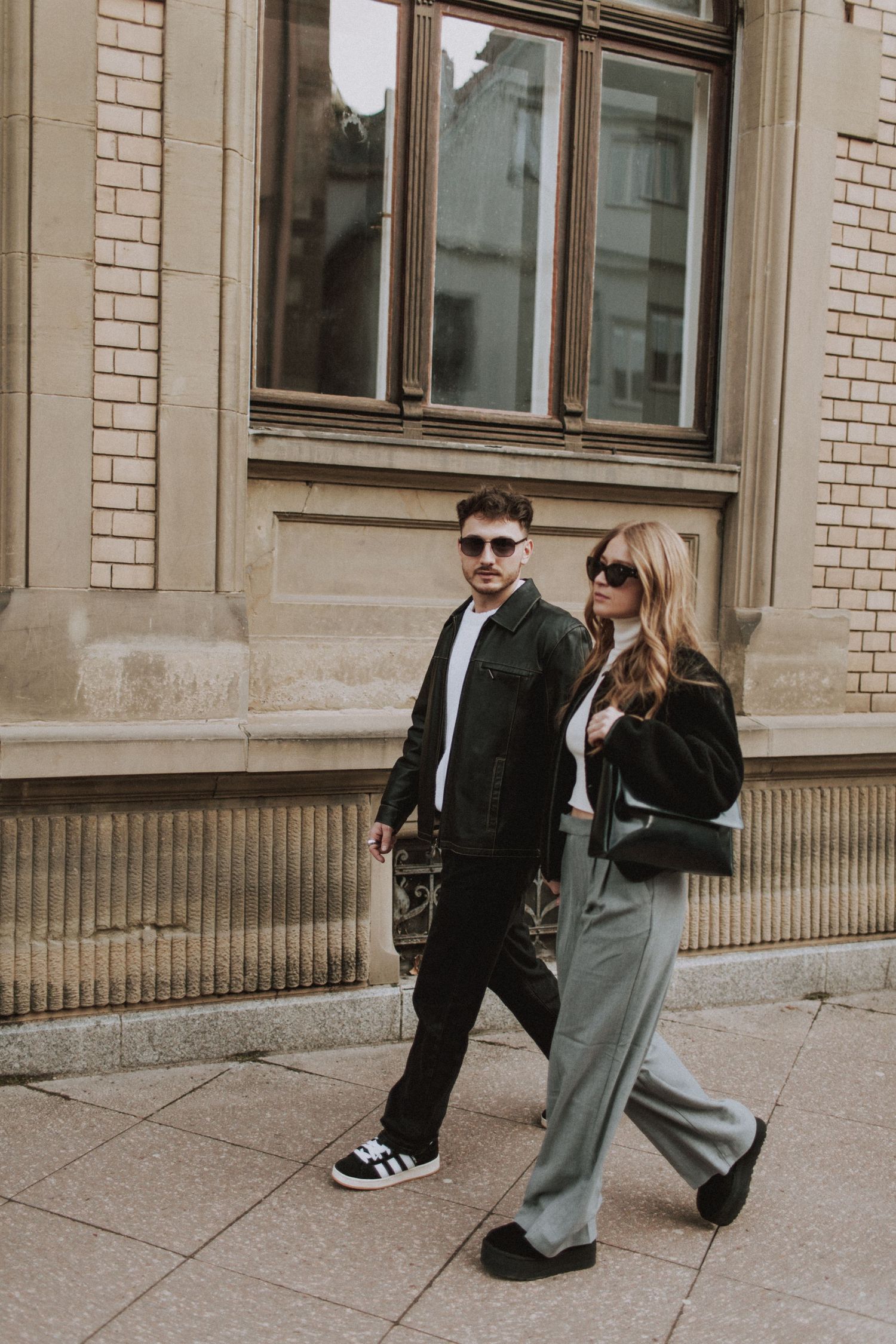 A fashionable couple walking together on a city sidewalk in casual urban attire.