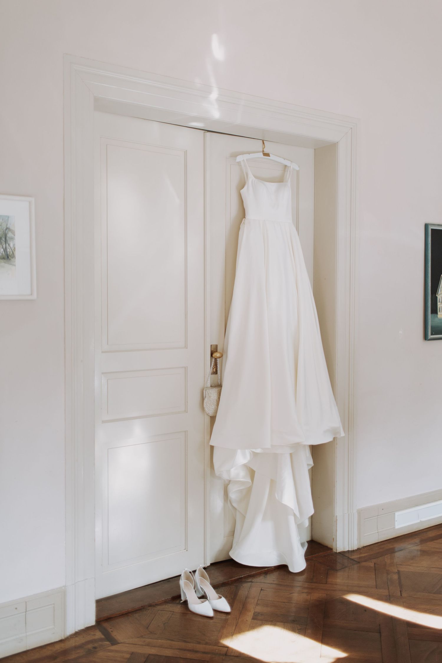 White wedding dress hanging on white door in sunlit room.