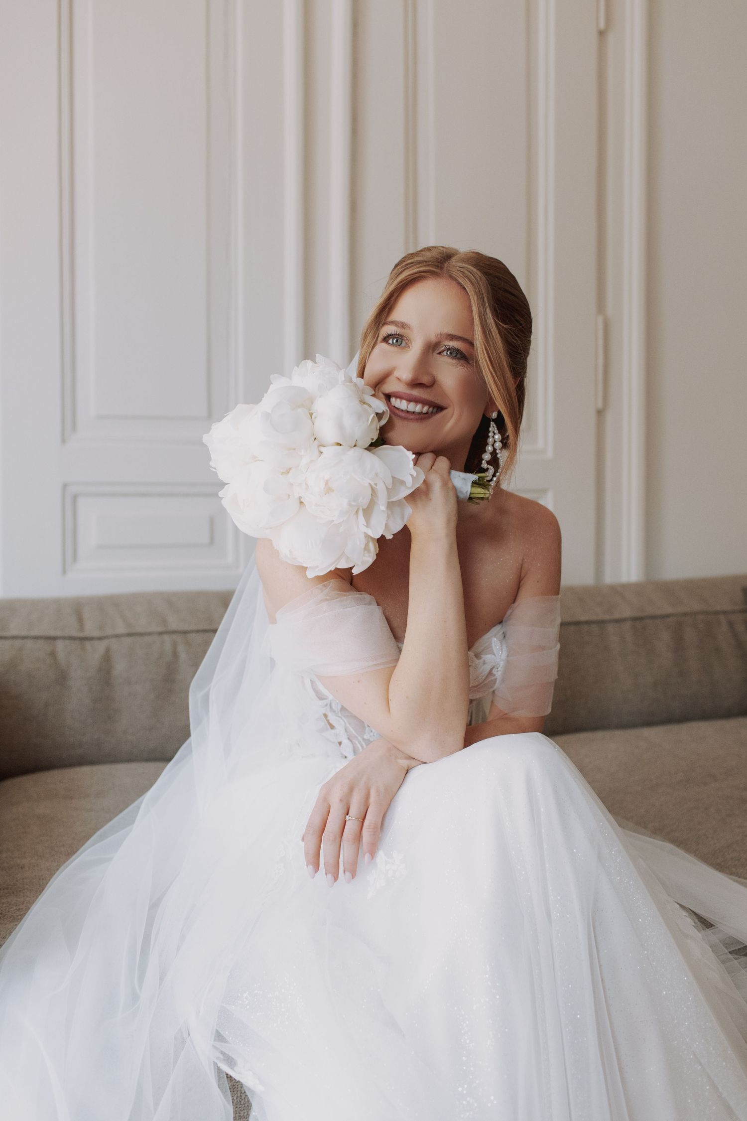 A joyful bride in a white tulle gown holds a bouquet of white peonies while sitting on a gray couch.