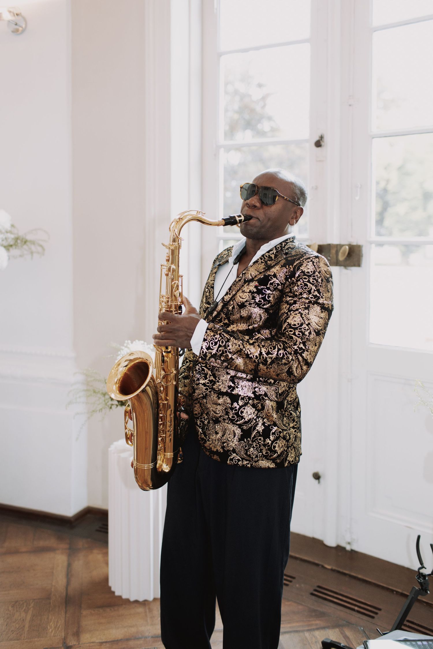 A musician in a floral brocade jacket plays saxophone in an elegant white room.
