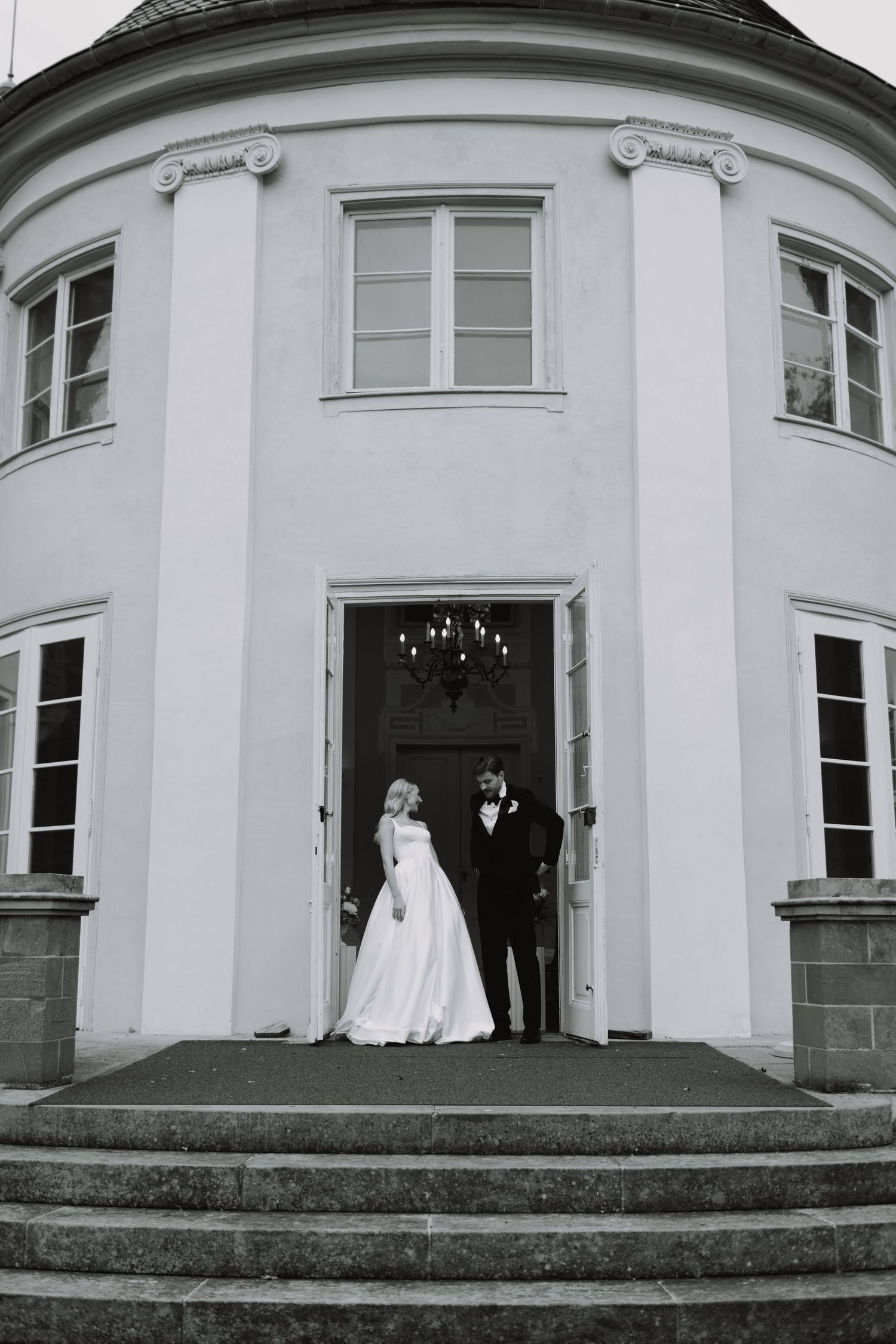 A couple stands in the doorway of an elegant white mansion with curved walls and classical architectural details.