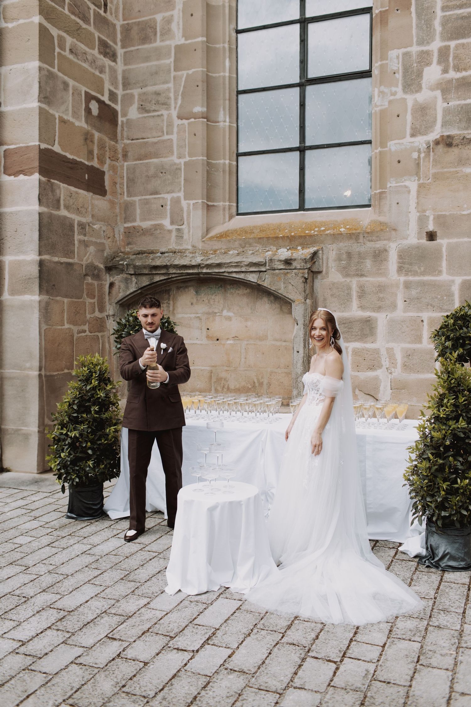 A wedding couple stands outside a stone church with elegant decor and potted topiaries.